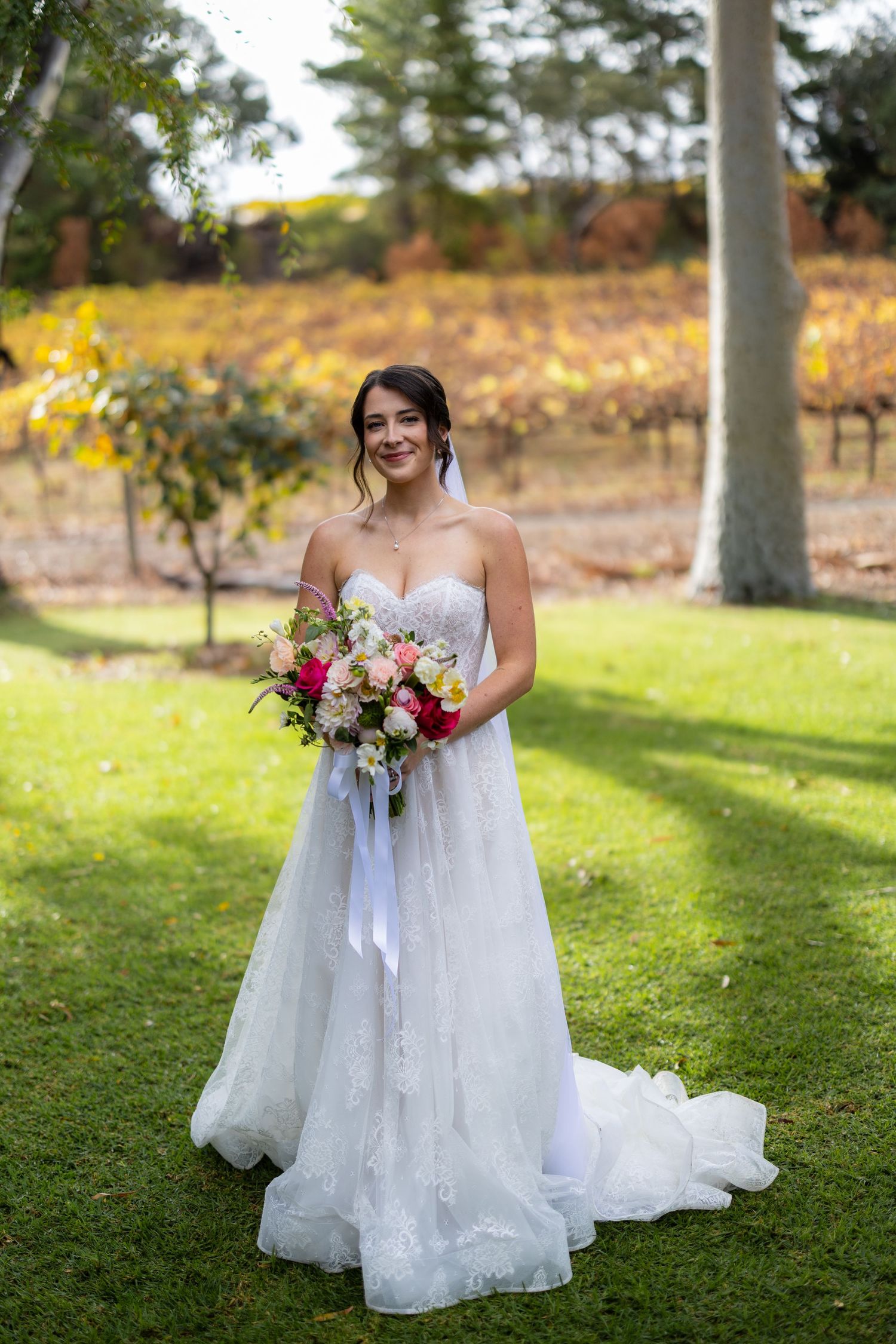 Elegant bride in strapless lace ball gown holds colorful bouquet while standing in front of autumn vineyard.
