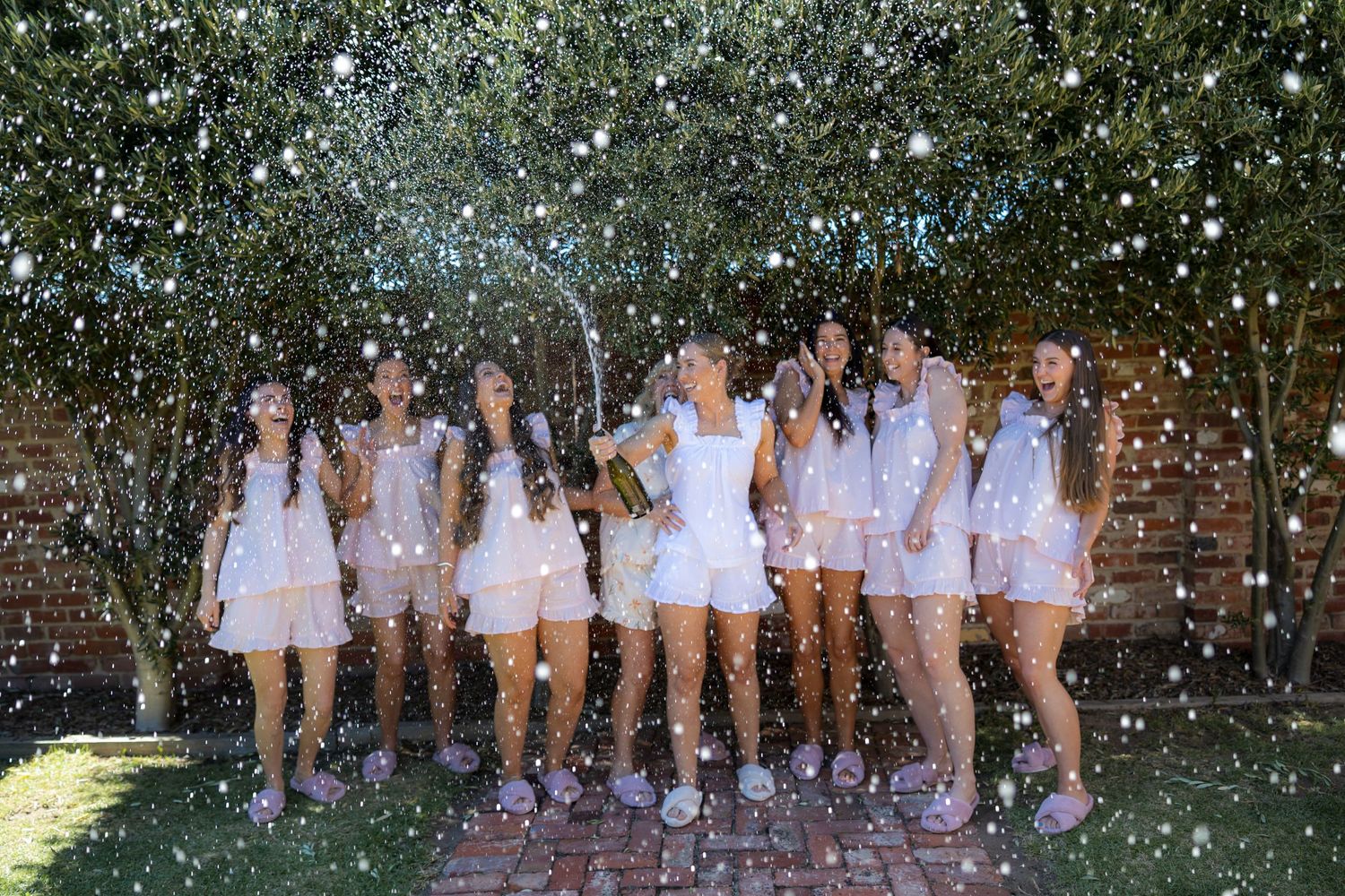 Bridal party in matching white outfits celebrating with champagne spray in garden setting.