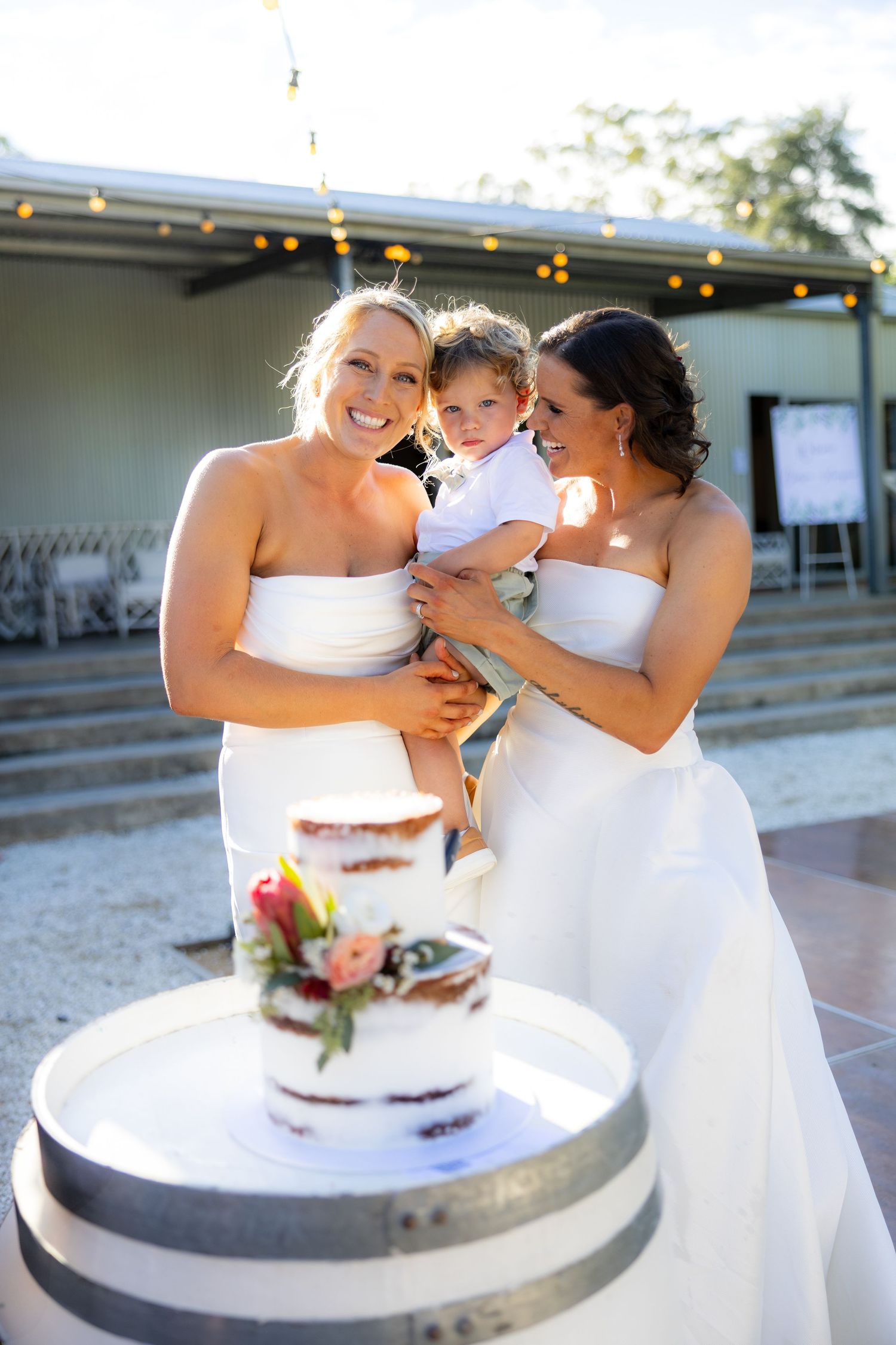 Two brides in white strapless gowns pose with a young child beside a floral wedding cake on a barrel.
