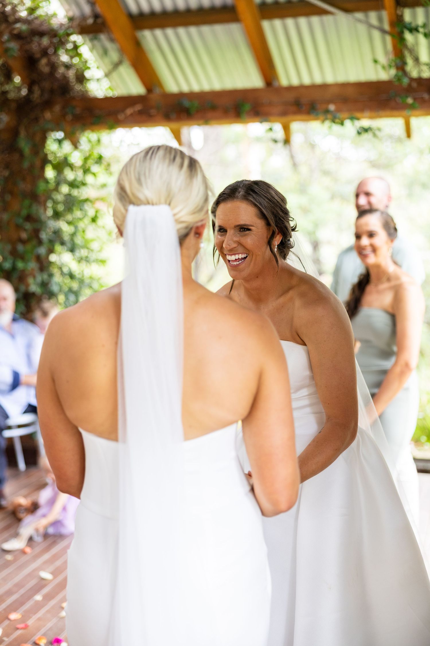 Two brides in white strapless gowns share a joyful moment during their wedding ceremony under a rustic pergola.