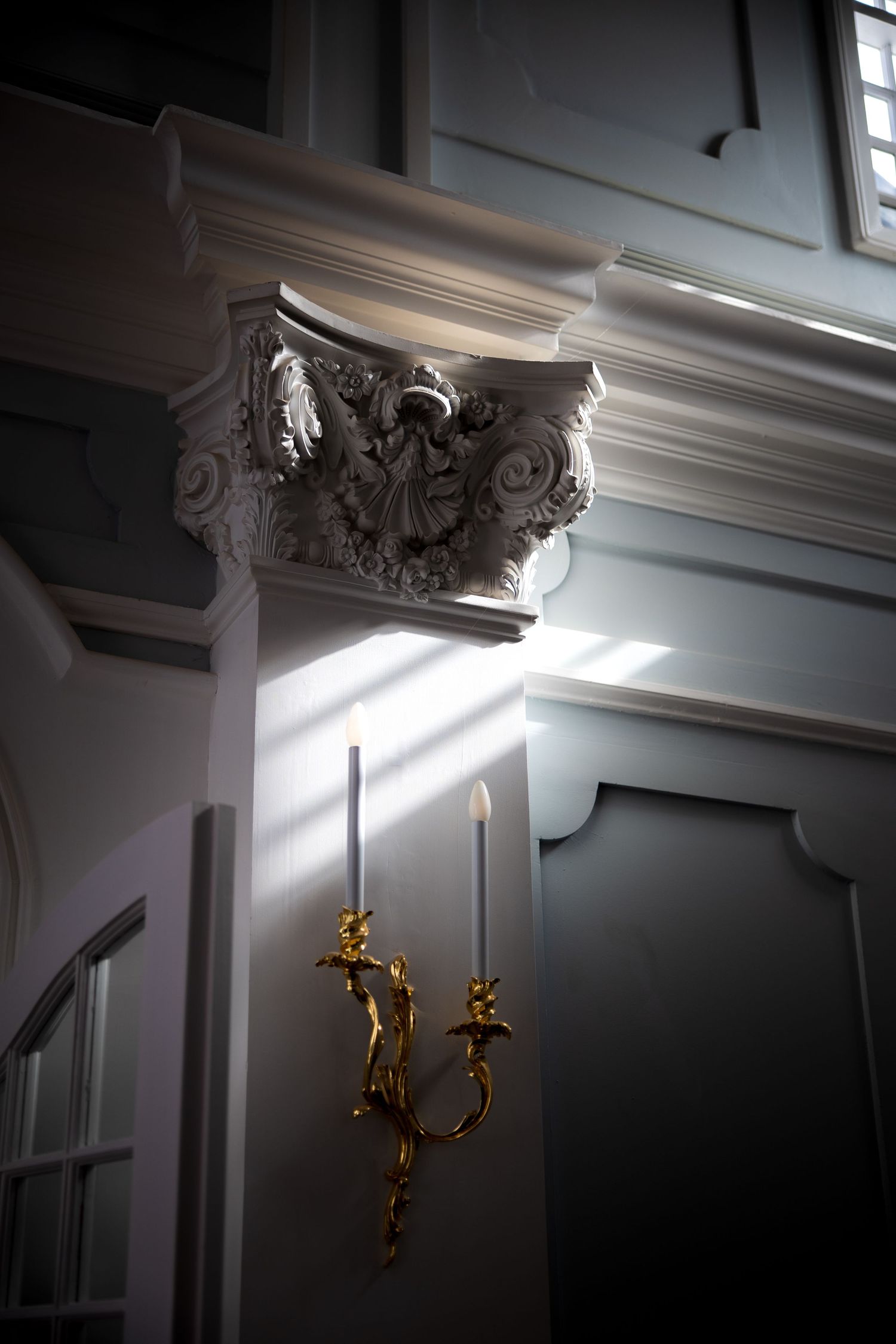 Ornate baroque column capital with gold candelabra wall sconce illuminated by dramatic light rays.