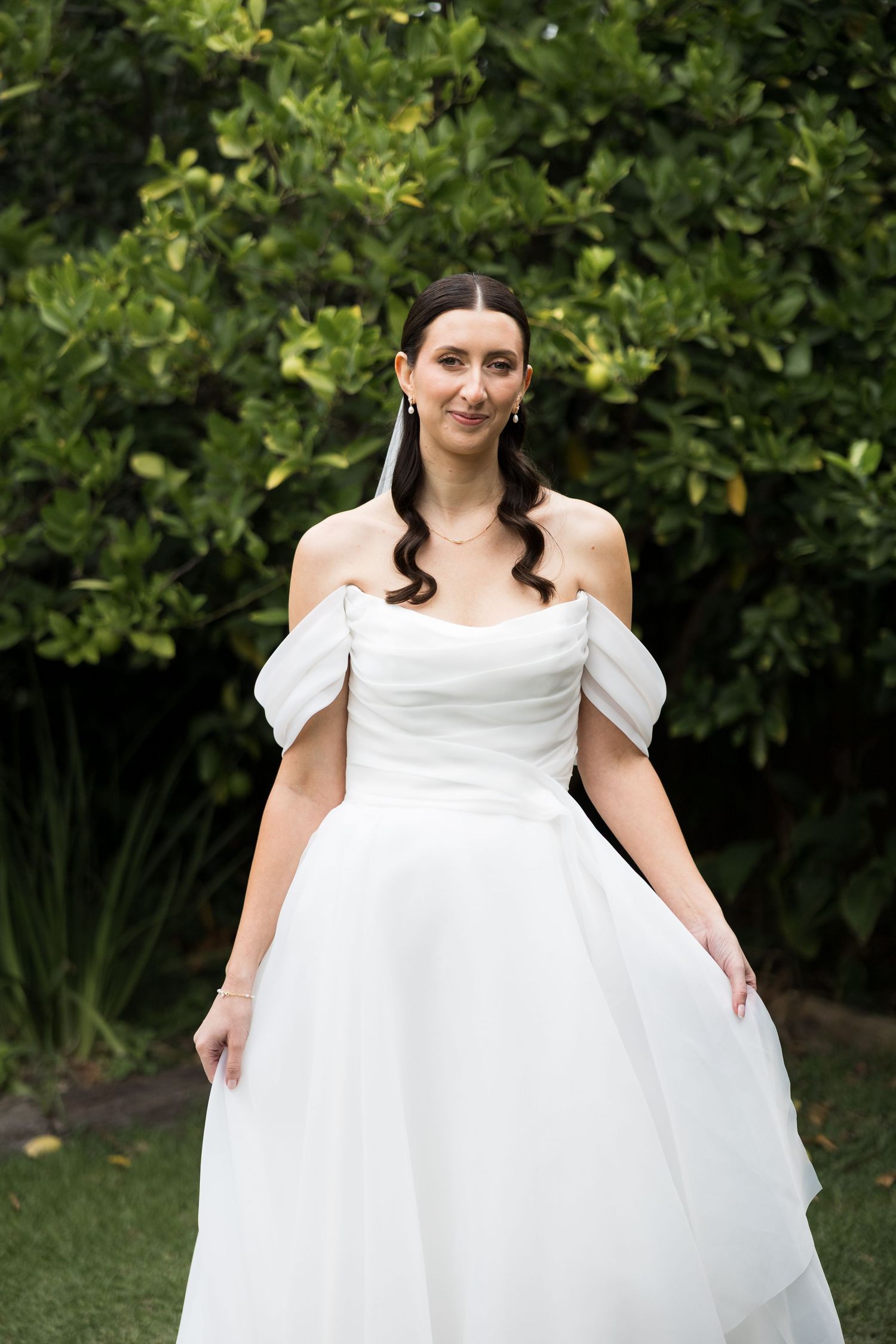 Bride in off-shoulder white ball gown poses outdoors against lush green foliage, with elegant curled hair.
