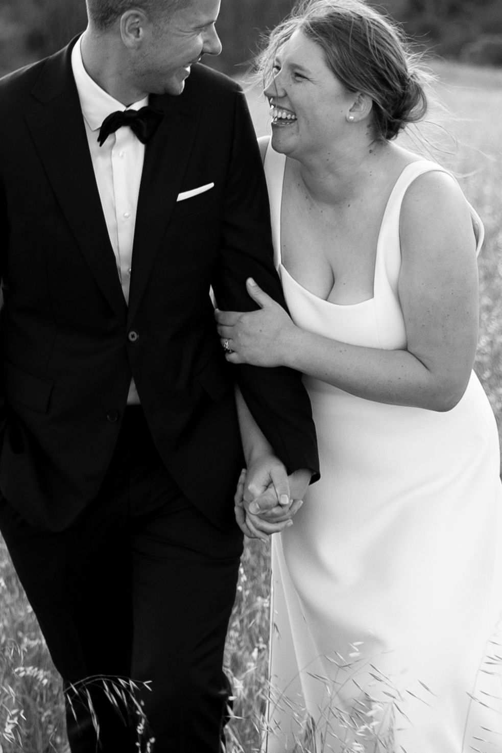 Black and white photo of a laughing bride and groom holding hands in an open field.