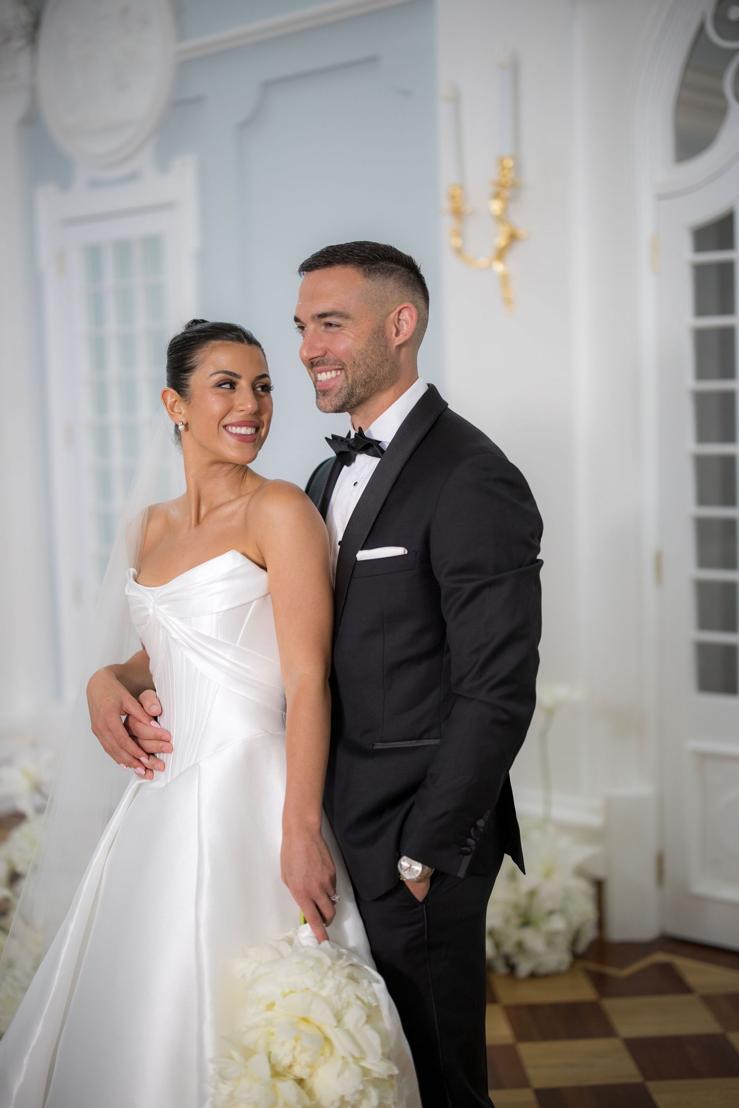 Smiling bride in strapless white gown and groom in black tuxedo posing together in elegant blue venue.