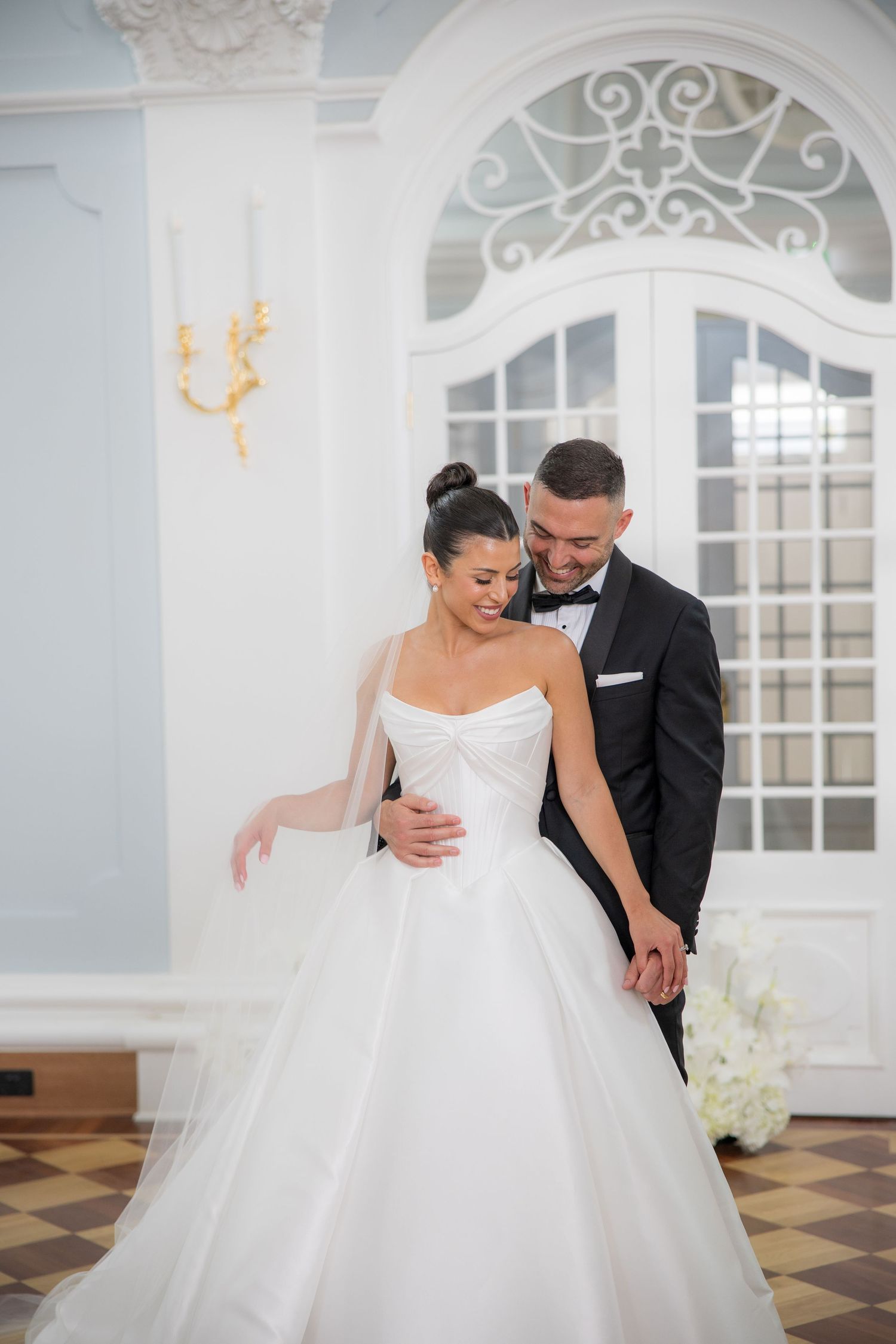 Smiling bride and groom share tender moment in elegant blue and white ballroom, groom in black tuxedo.