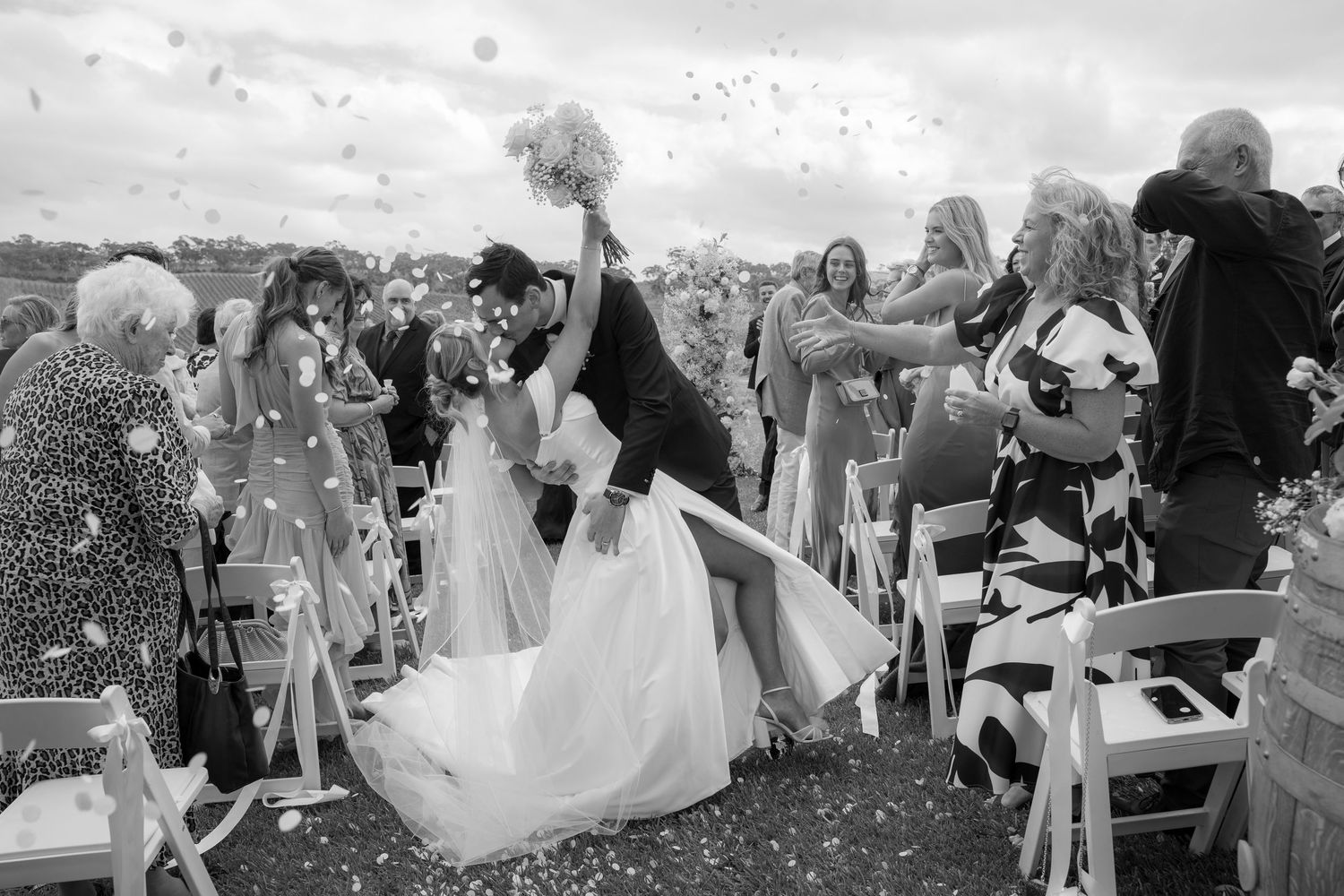 Black and white photo of groom dipping bride for kiss during confetti exit at wedding ceremony.
