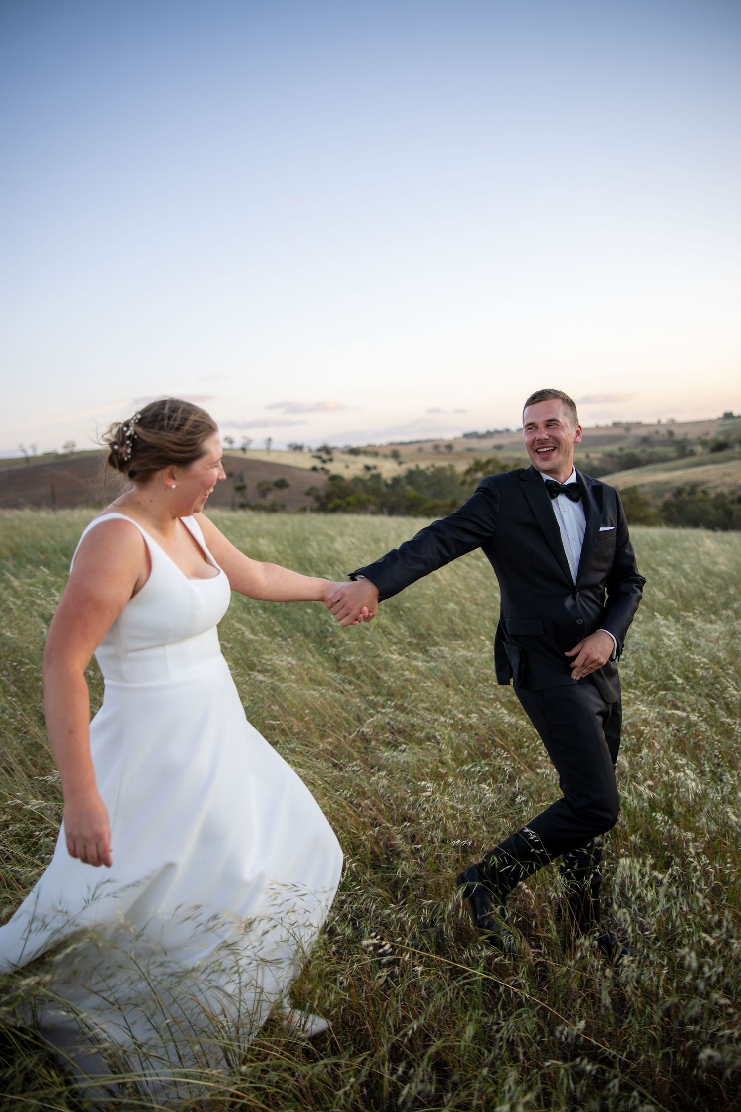 Joyful bride and groom holding hands and laughing in a scenic green field at sunset.