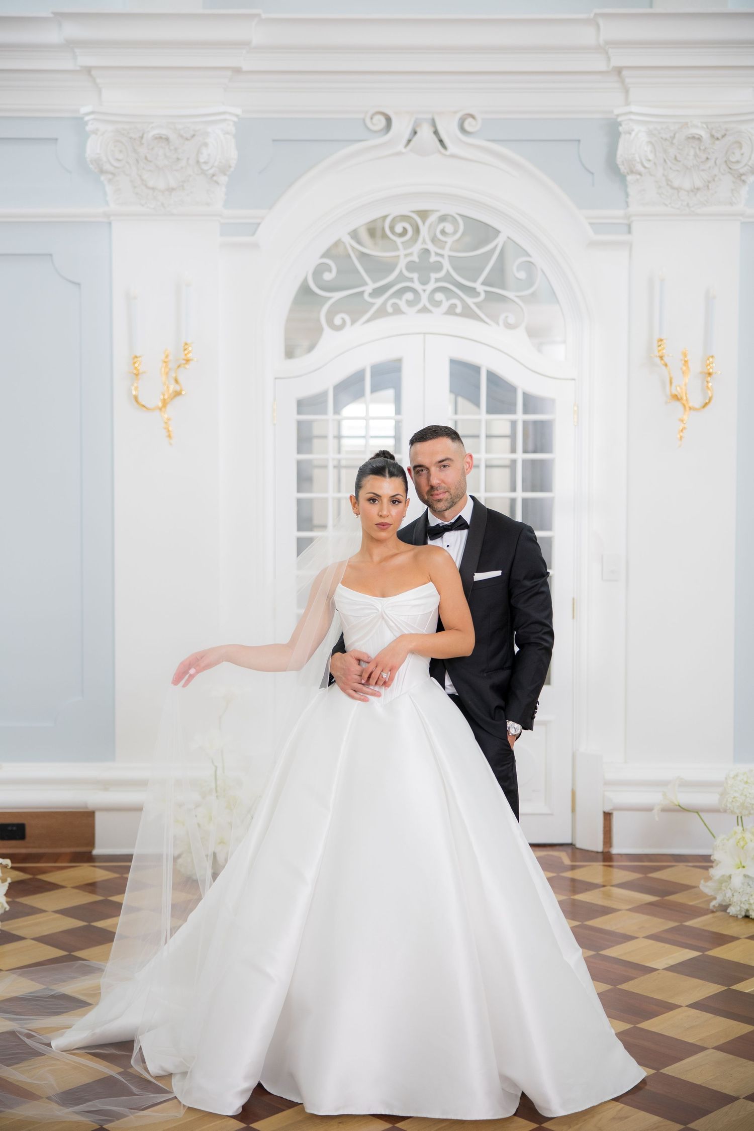 Elegant bride in white ballgown and groom in black tuxedo posing in a grand white ornate ballroom.