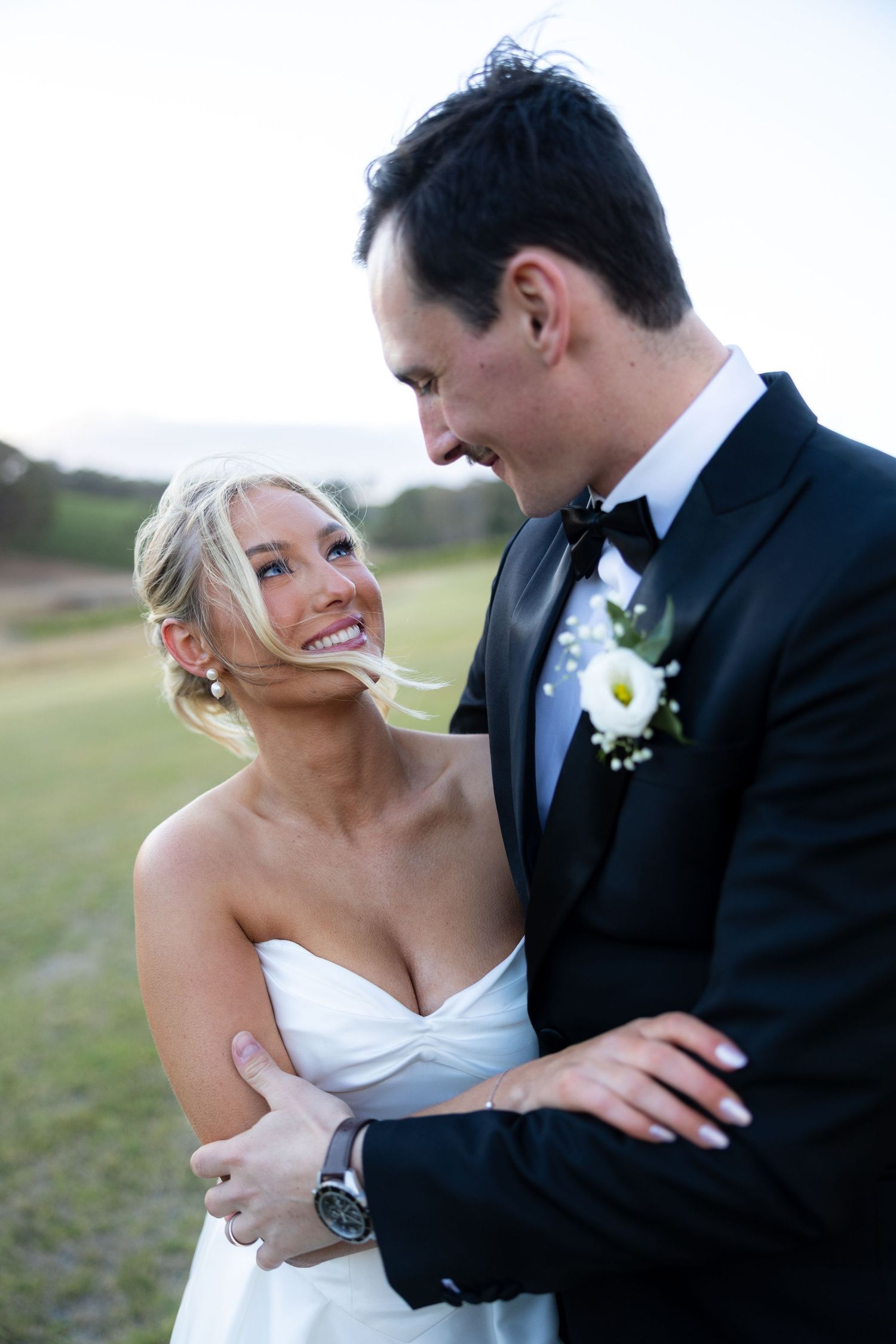 Smiling blonde bride in strapless white gown embraced by groom in black tuxedo with boutonniere outdoors.