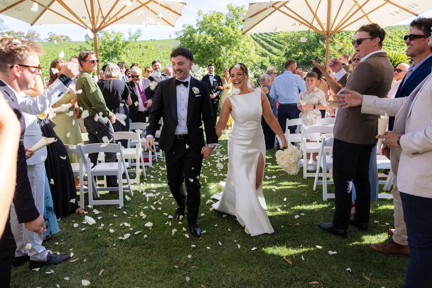 Bride and groom walking down aisle as guests toss white petals at sunny outdoor vineyard wedding.