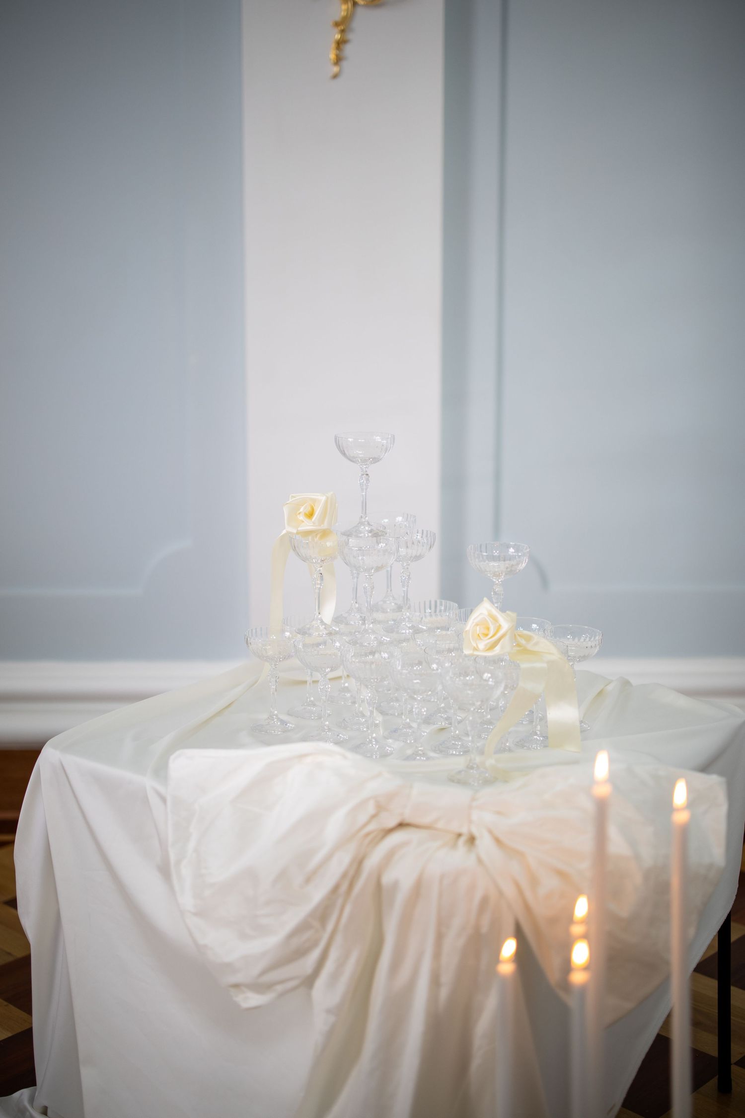 Wedding champagne tower with crystal glasses arranged on a white draped table with lit candles and cream roses.