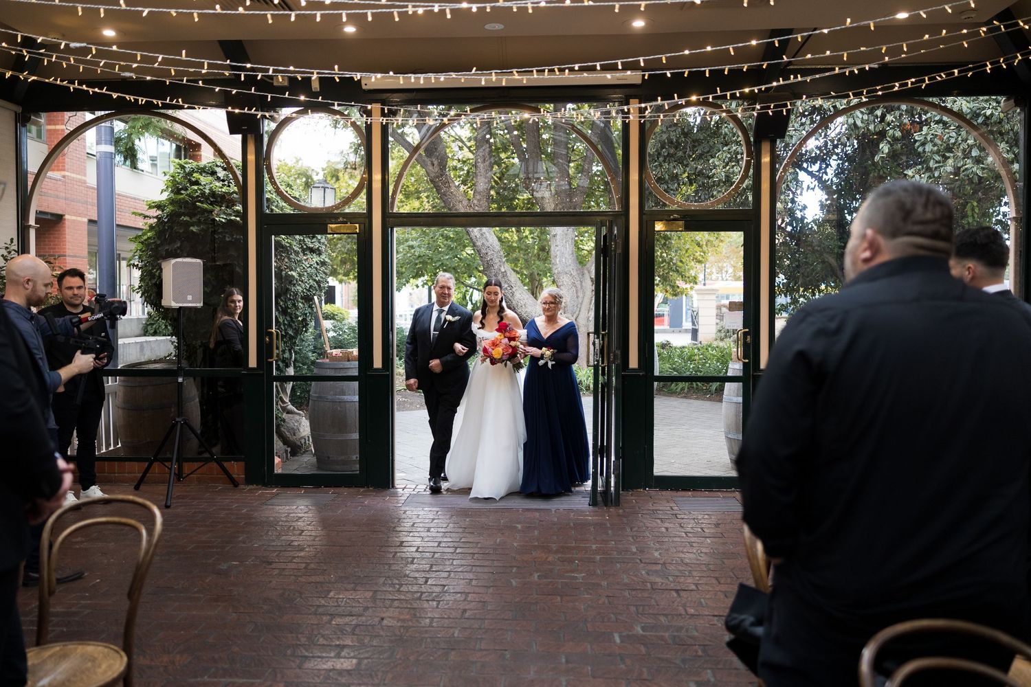 Bride and groom stand at outdoor wedding ceremony entrance under string lights at elegant brick courtyard venue.