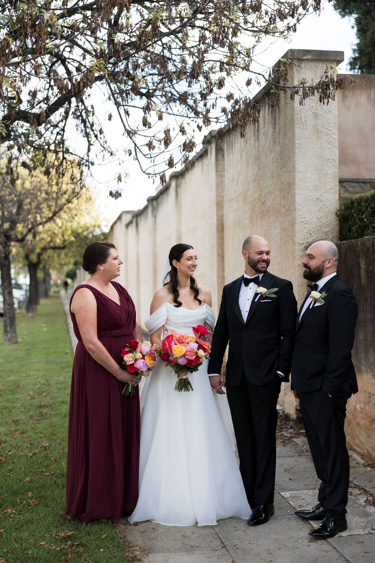 Wedding party of four posing outdoors, bride in white gown with bouquet, bridesmaid in burgundy, two grooms in black tuxedos.