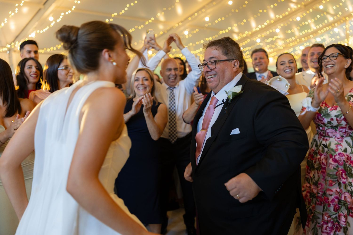 Joyful wedding reception guests cheer as bride in white gown dances with smiling man in suit.