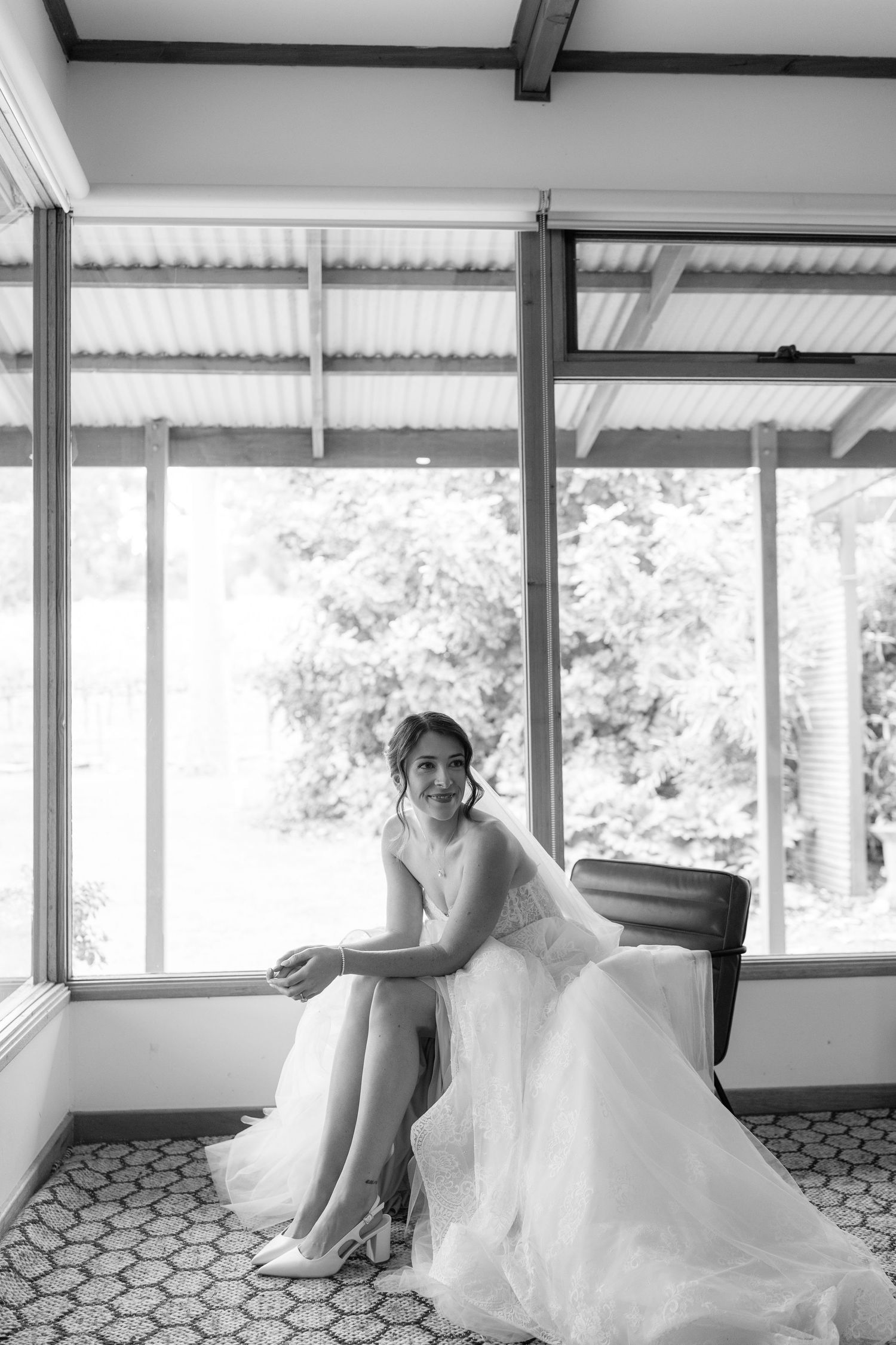 Elegant bride in white tulle gown sits by large windows in black and white bridal portrait photo.