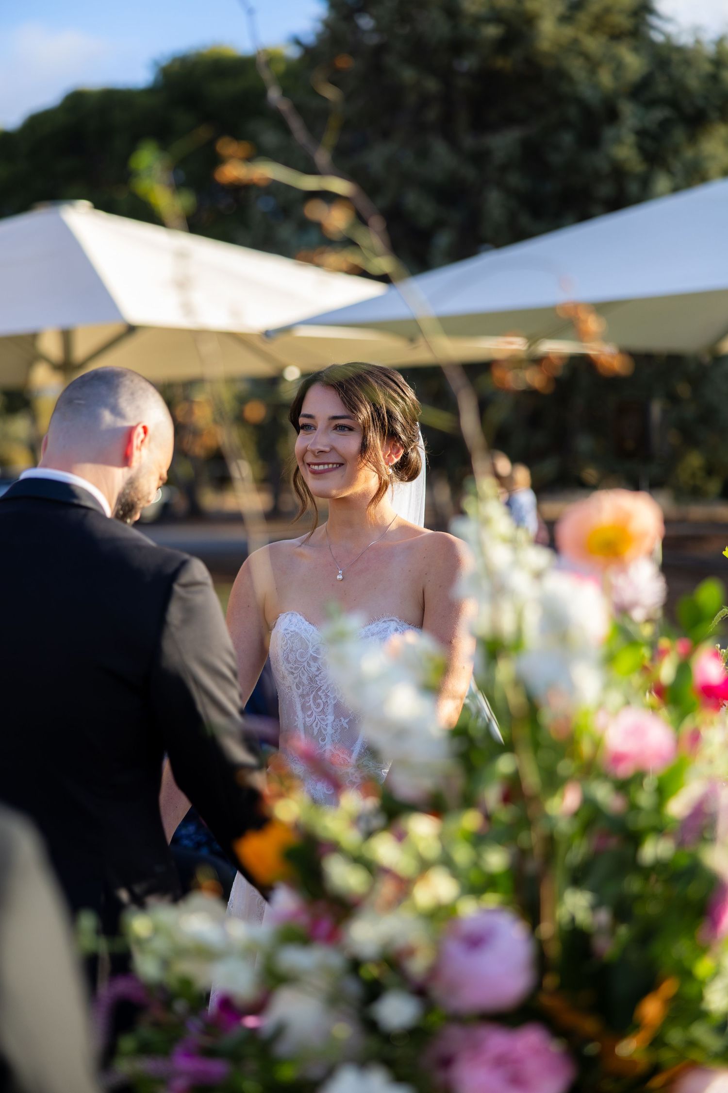 Smiling bride in strapless lace gown holds hands with groom during outdoor wedding ceremony surrounded by colorful florals.