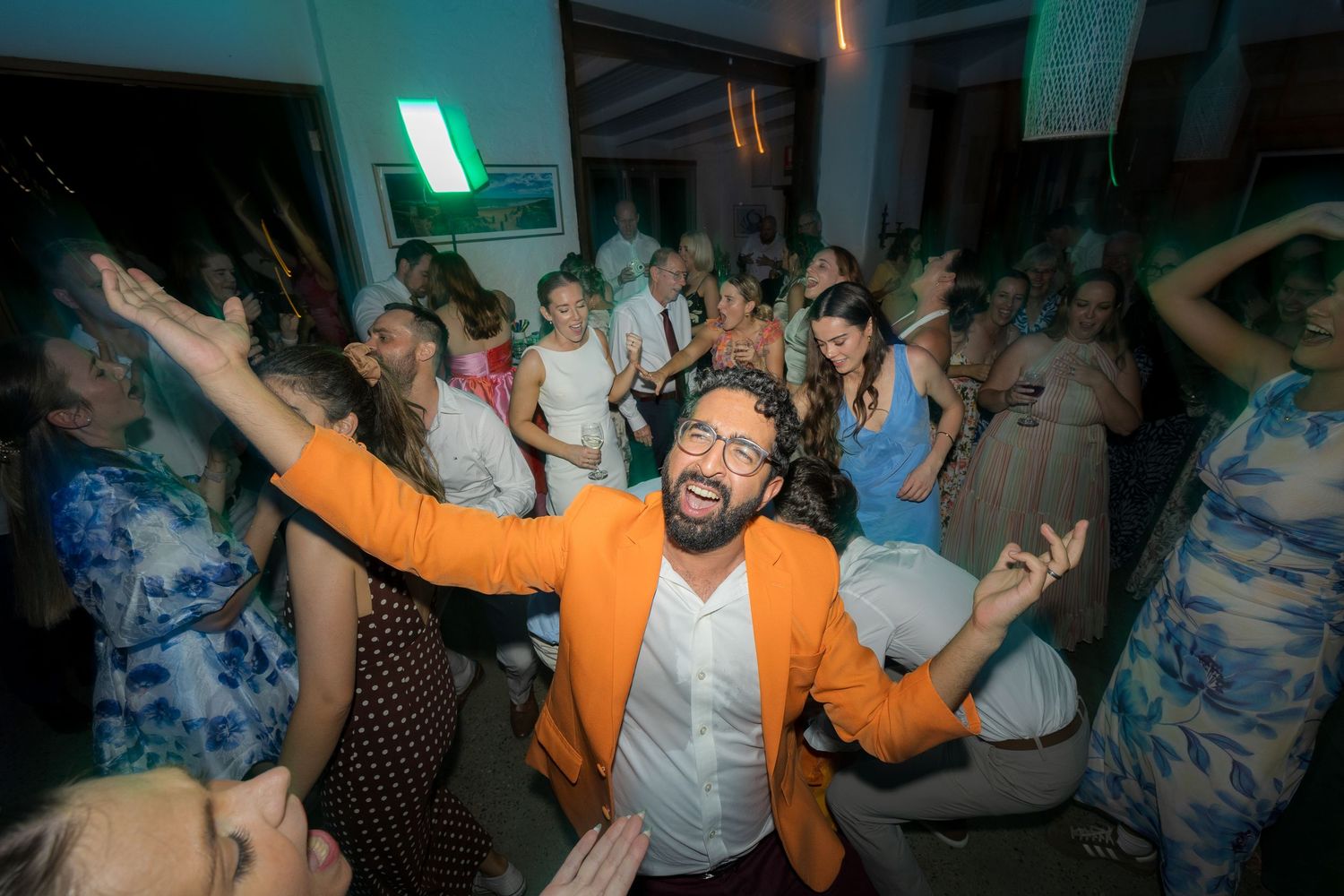 Man in orange blazer dances energetically with a large crowd of guests at a lively wedding reception.
