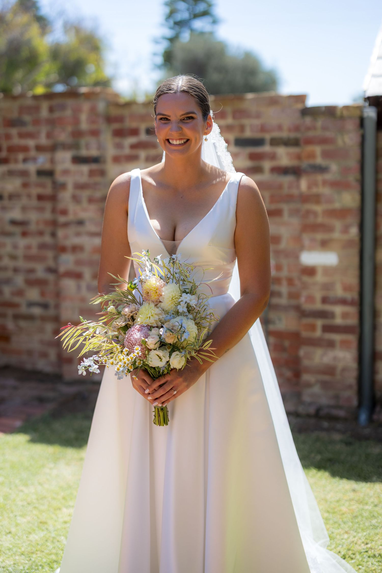 Smiling bride in V-neck wedding gown holding pastel wildflower bouquet in sunny garden setting.
