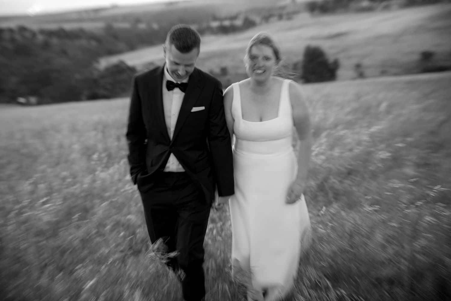 Black and white photo of a laughing bride and groom walking through a field on their wedding day.