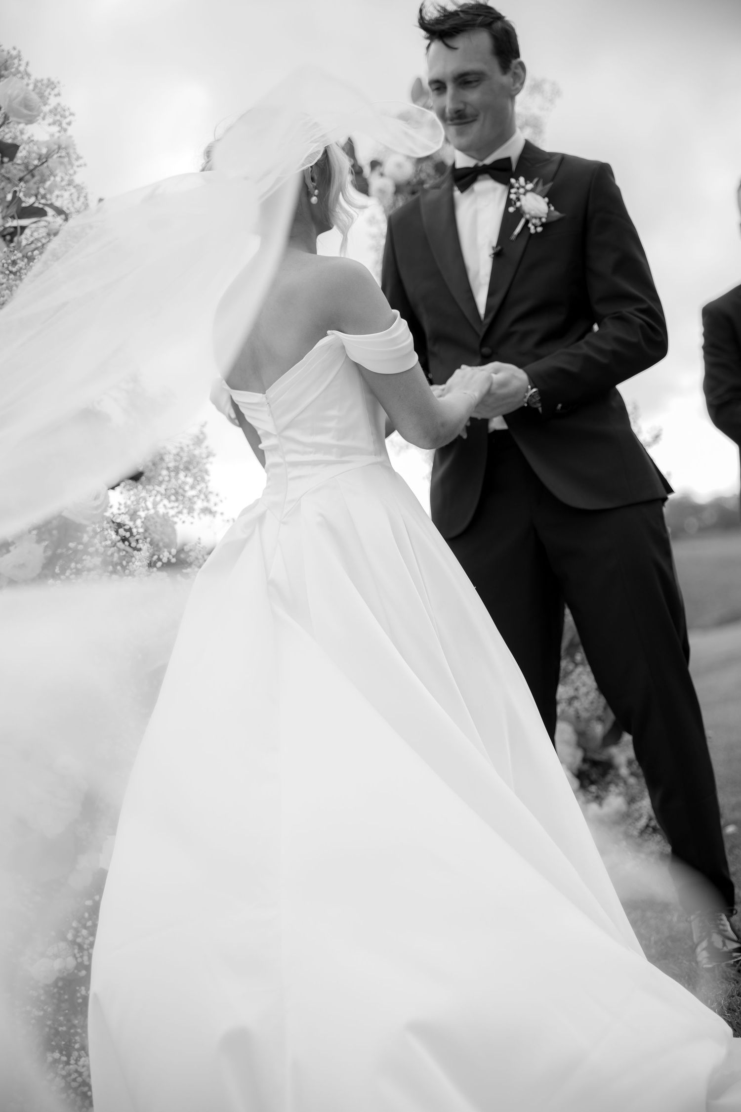 Black and white photo of bride and groom holding hands outdoors, veil flowing in the wind.