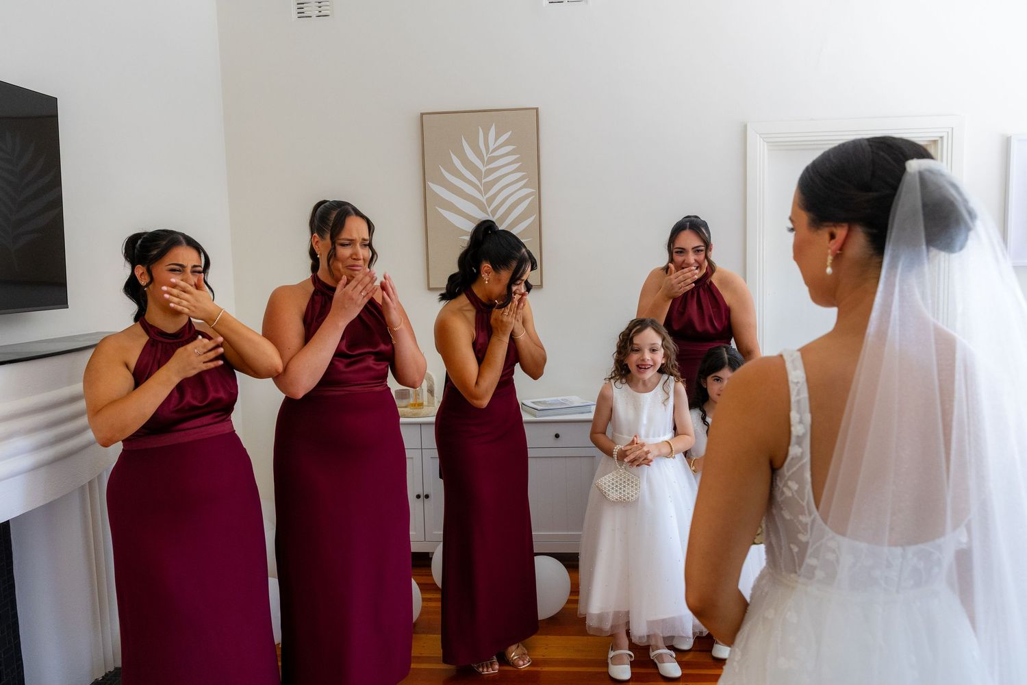 Bridesmaids in burgundy dresses reacting emotionally to bride in white wedding gown and veil.