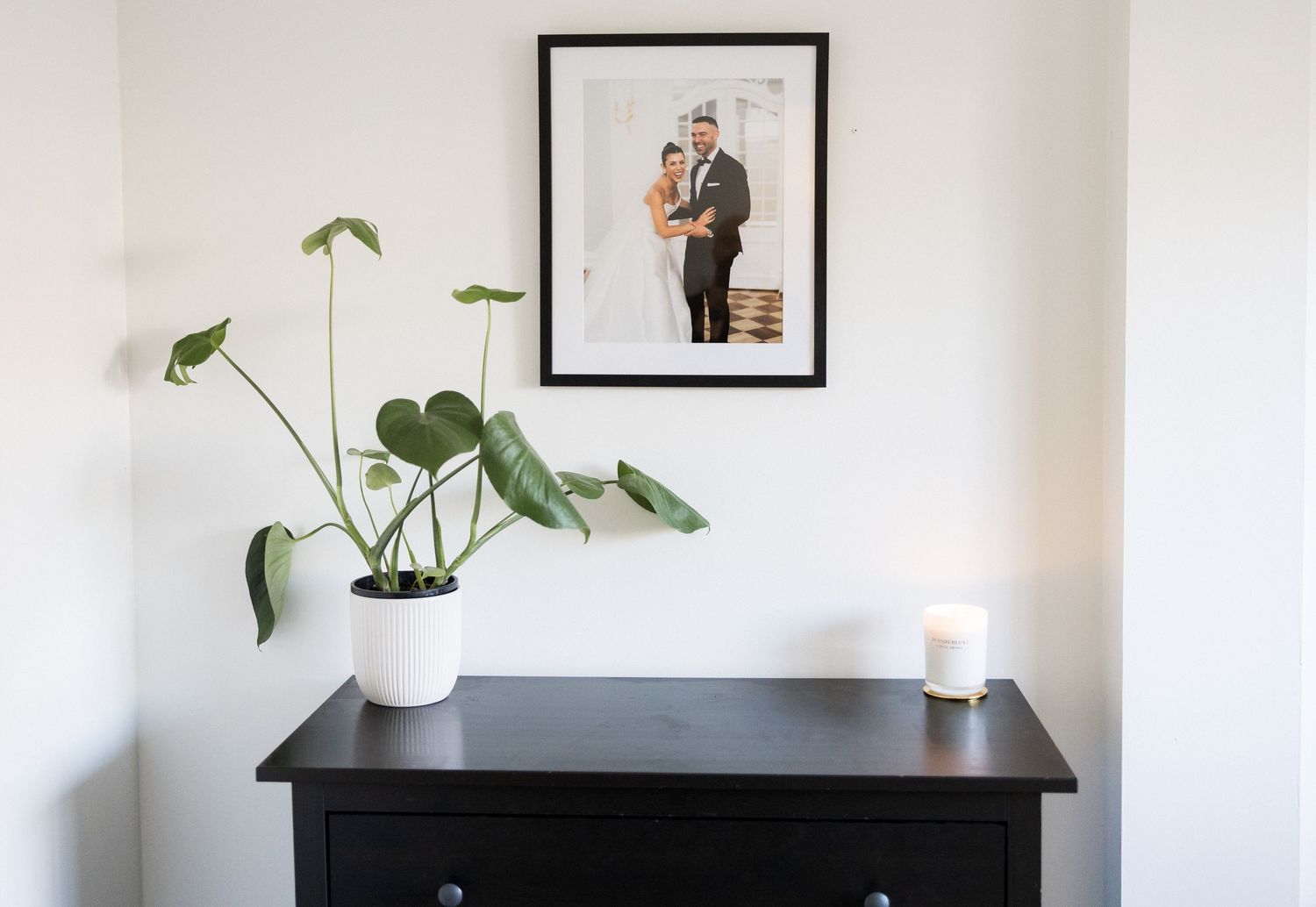 Wedding portrait in black frame above dark dresser with white potted monstera and candle.