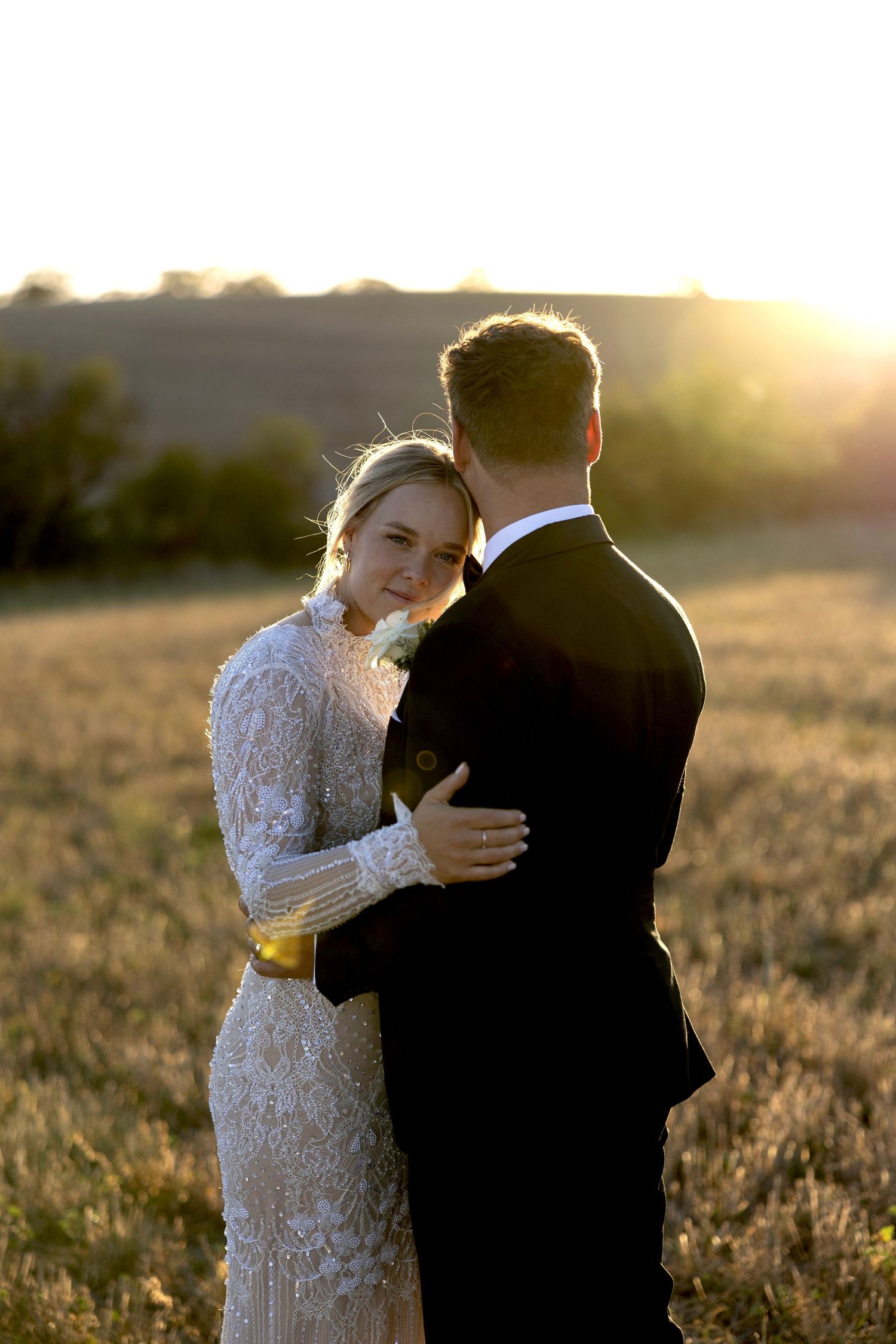 Bride and groom share a romantic embrace in a golden wheat field during a stunning sunset portrait.