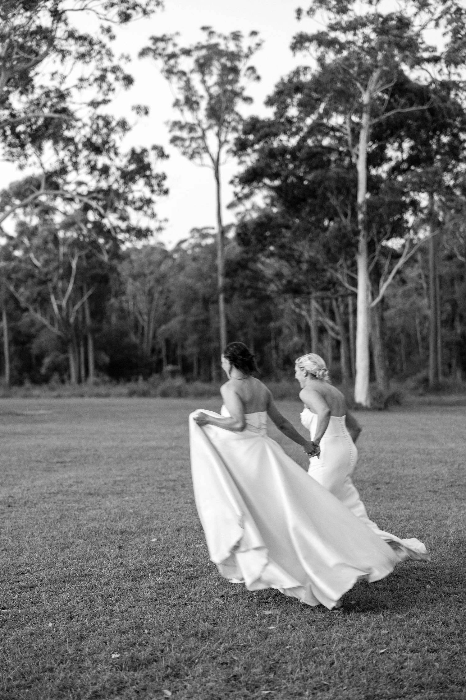 Black and white photo of two brides in white gowns walking hand in hand across an open field.
