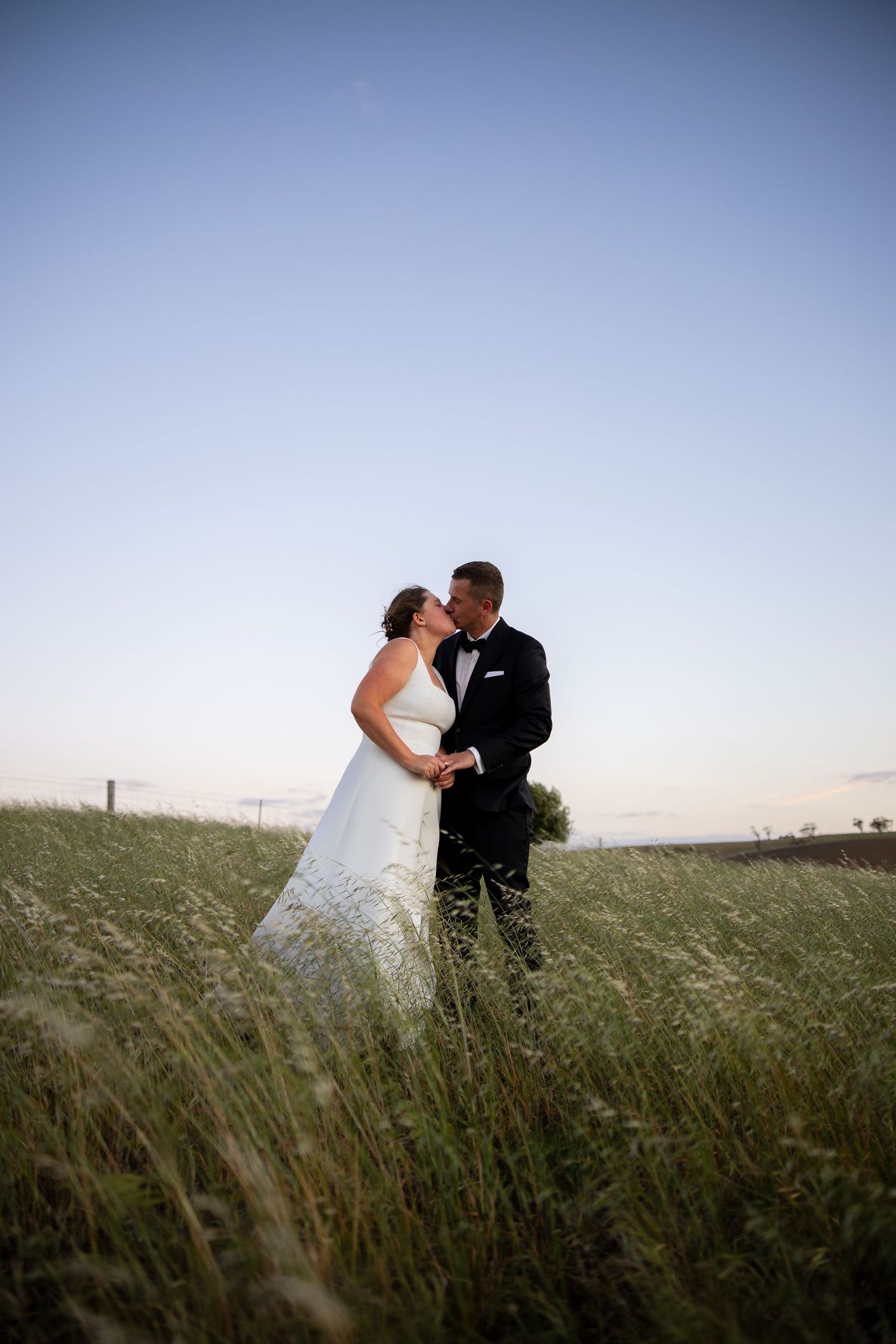 Bride and groom share a romantic kiss in a lush green field at dusk, with a clear blue sky backdrop.