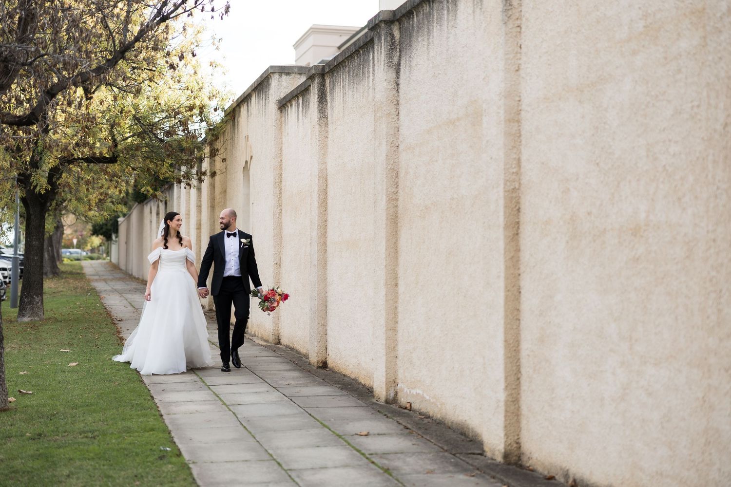 Bride and groom walking hand in hand along a tree-lined path beside a beige stone wall on their wedding day.