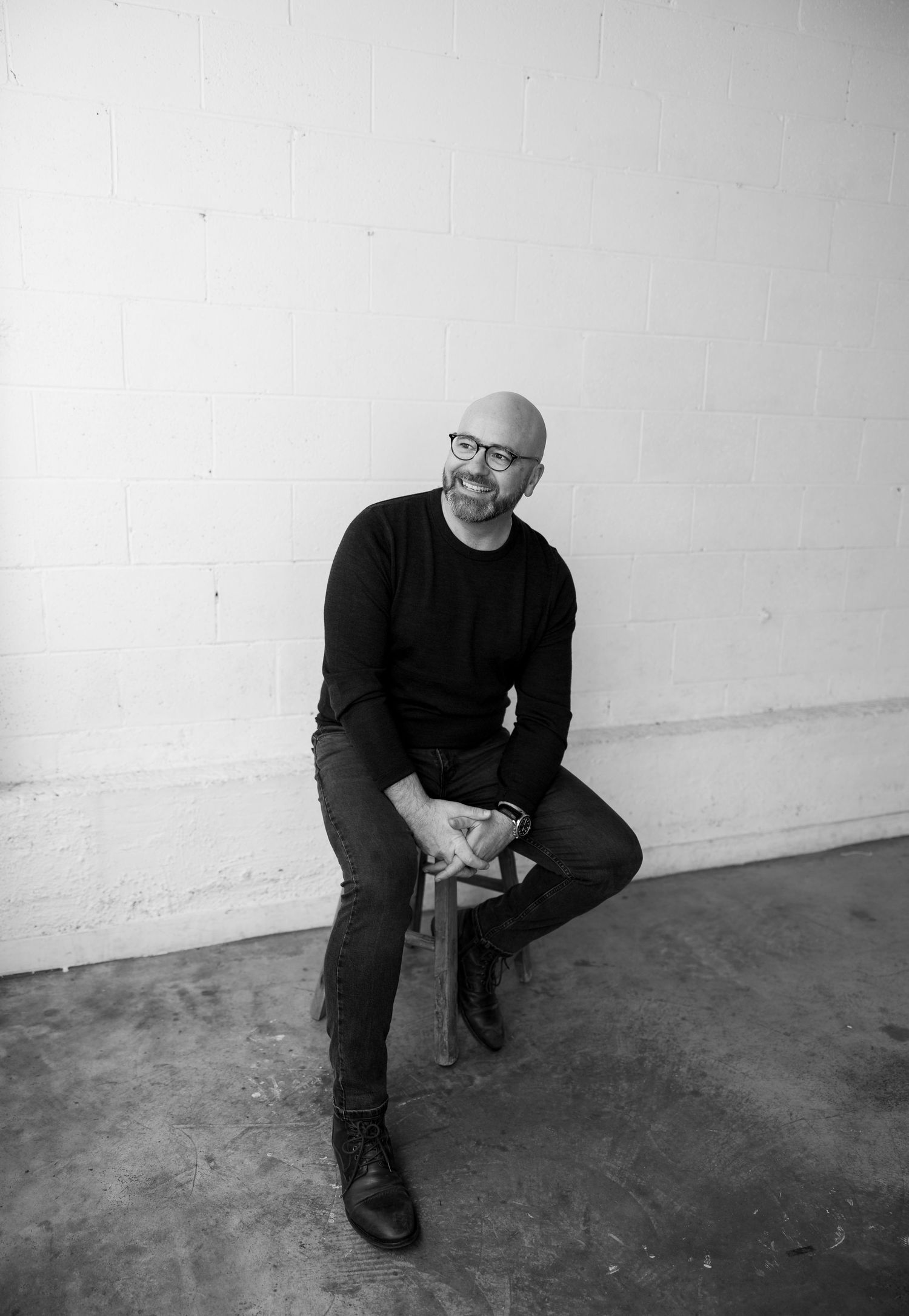 Bald man with glasses smiling, seated on stool in black outfit, black and white portrait photo.