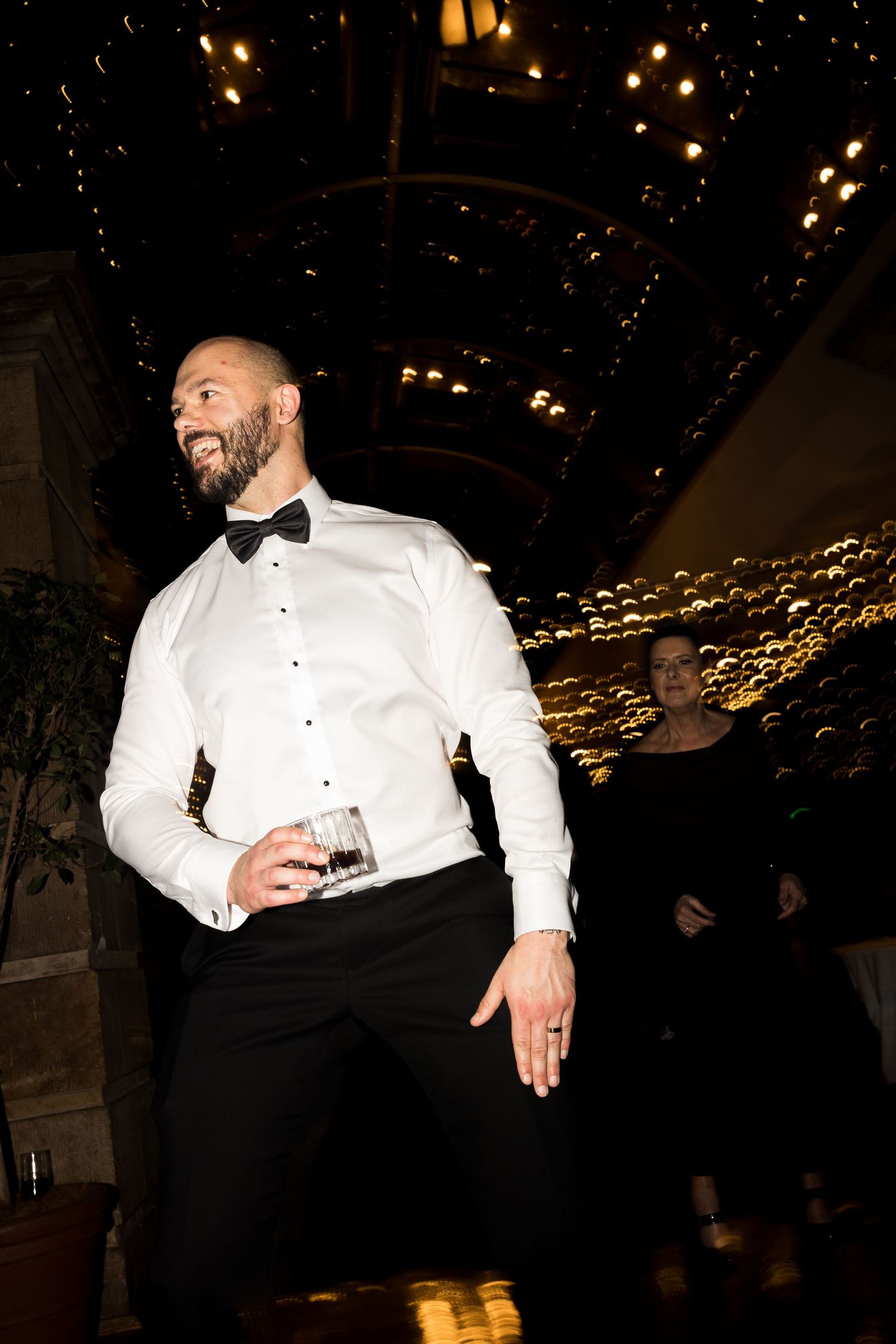 Smiling bald man in white tuxedo shirt and black bow tie holding a drink at an elegant formal event.