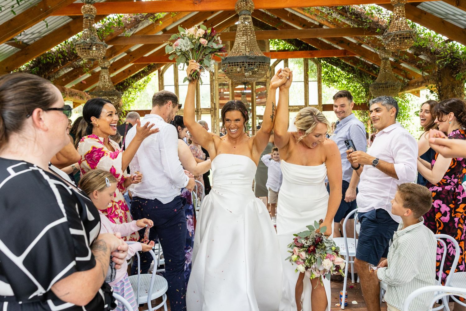 Two brides in white gowns celebrate joyfully with raised hands as guests cheer during their wedding reception exit.