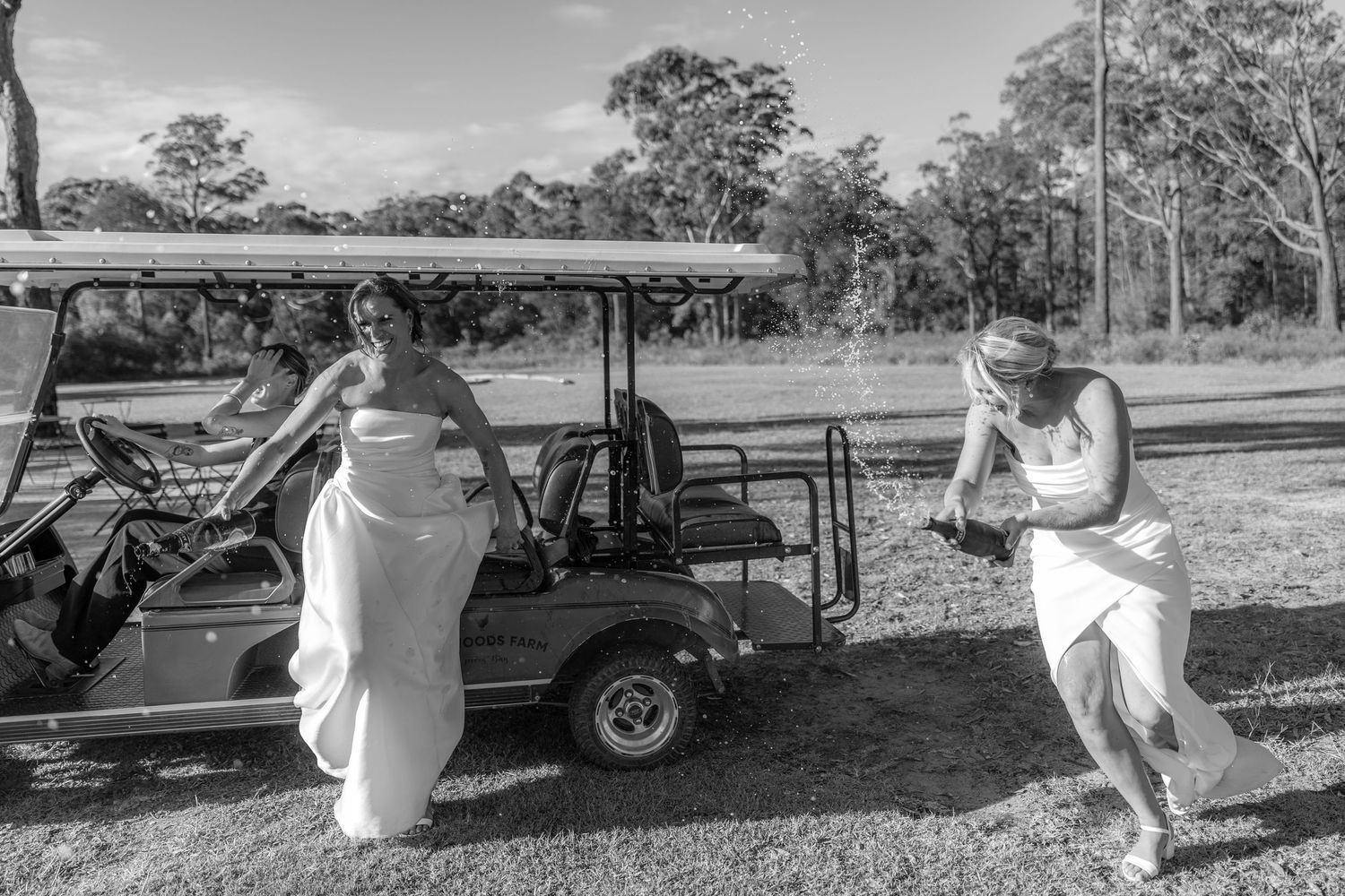 Black and white photo of two brides in white gowns near a golf cart at an outdoor wedding venue.
