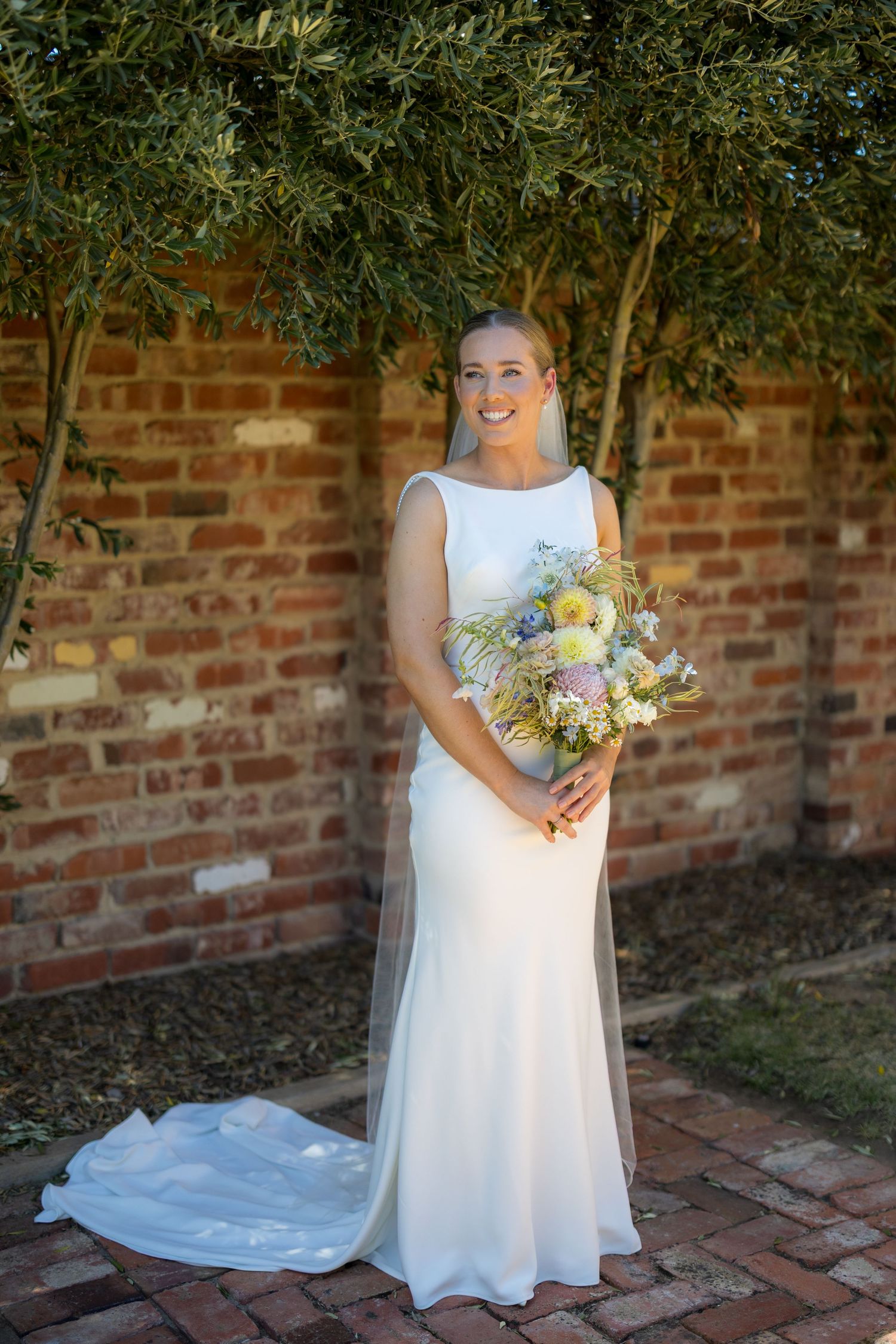 Smiling bride in white gown holding wildflower bouquet standing against brick wall with olive tree.