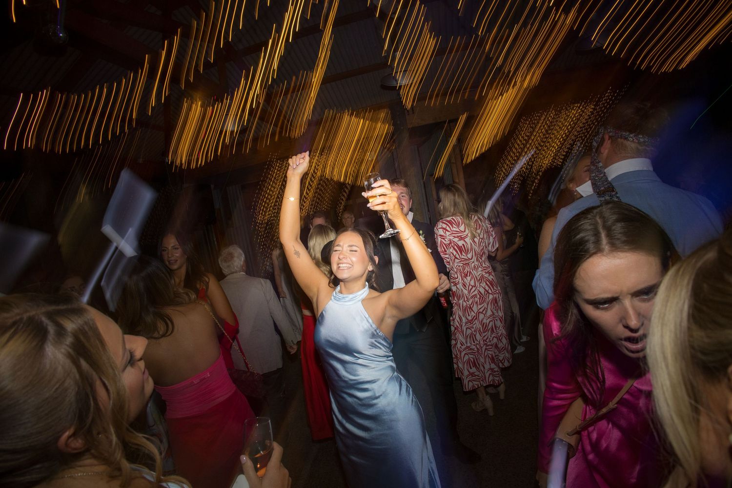 Wedding guest in blue satin dress dances joyfully at lively indoor wedding reception celebration.