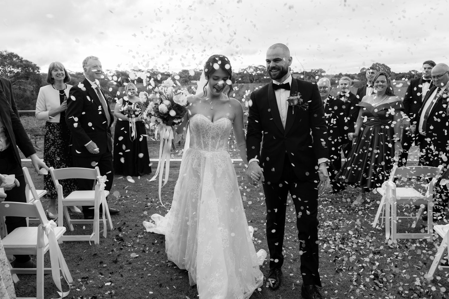 Black and white photo of newlyweds walking through confetti shower down outdoor wedding ceremony aisle.