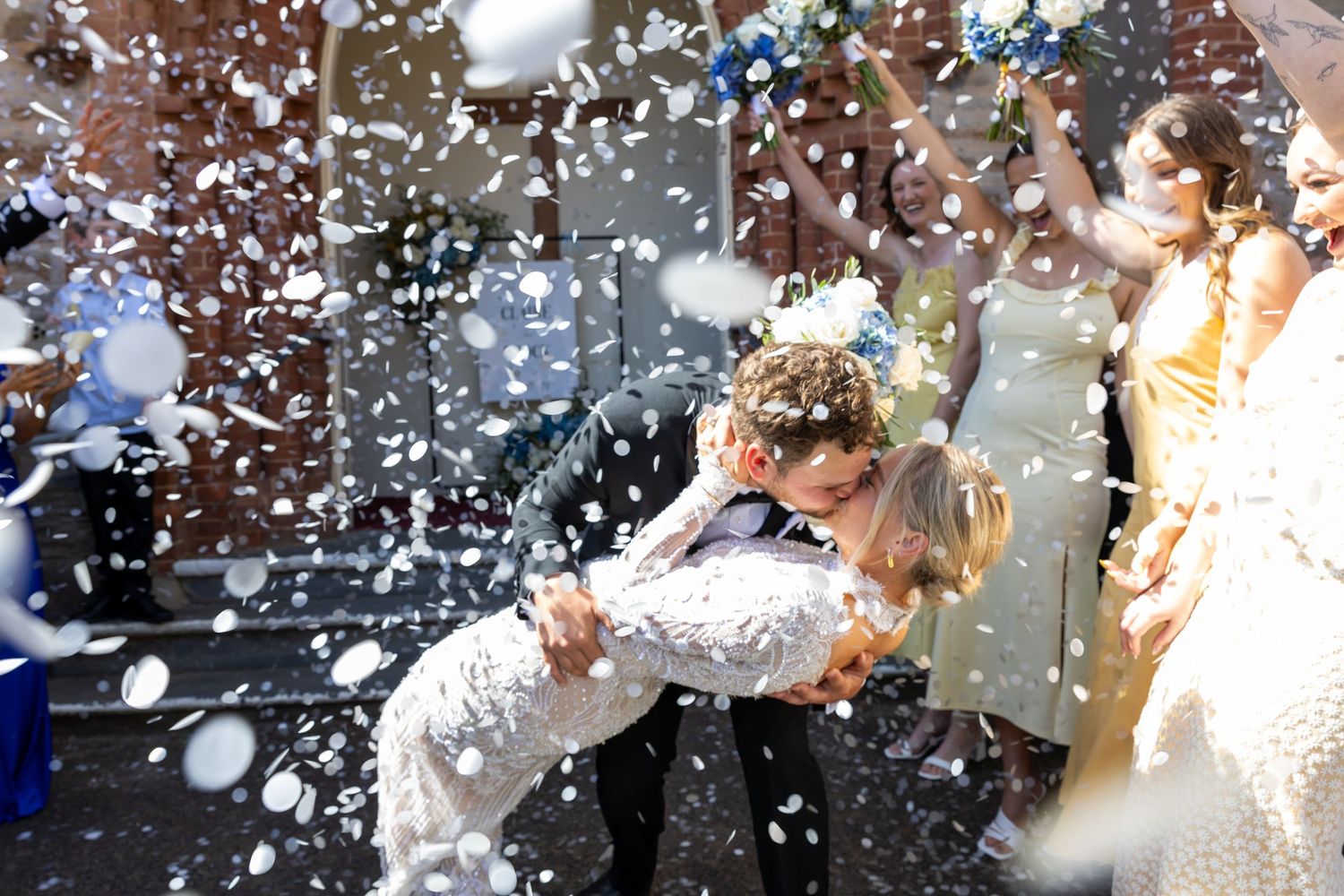 Newlyweds share a romantic dip kiss surrounded by guests throwing white confetti at their wedding celebration.