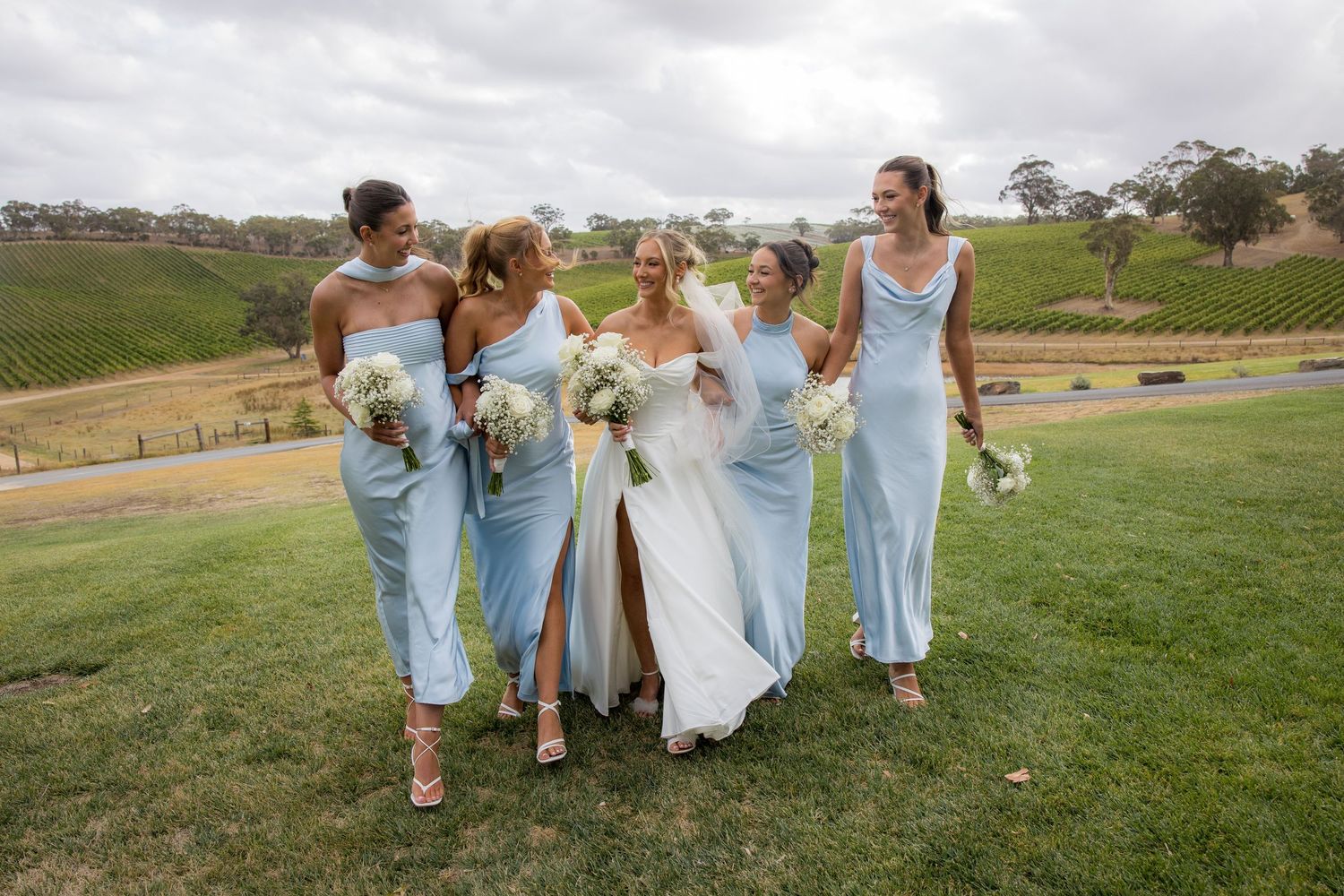 Bride and four bridesmaids in light blue satin dresses walk together laughing at vineyard wedding.