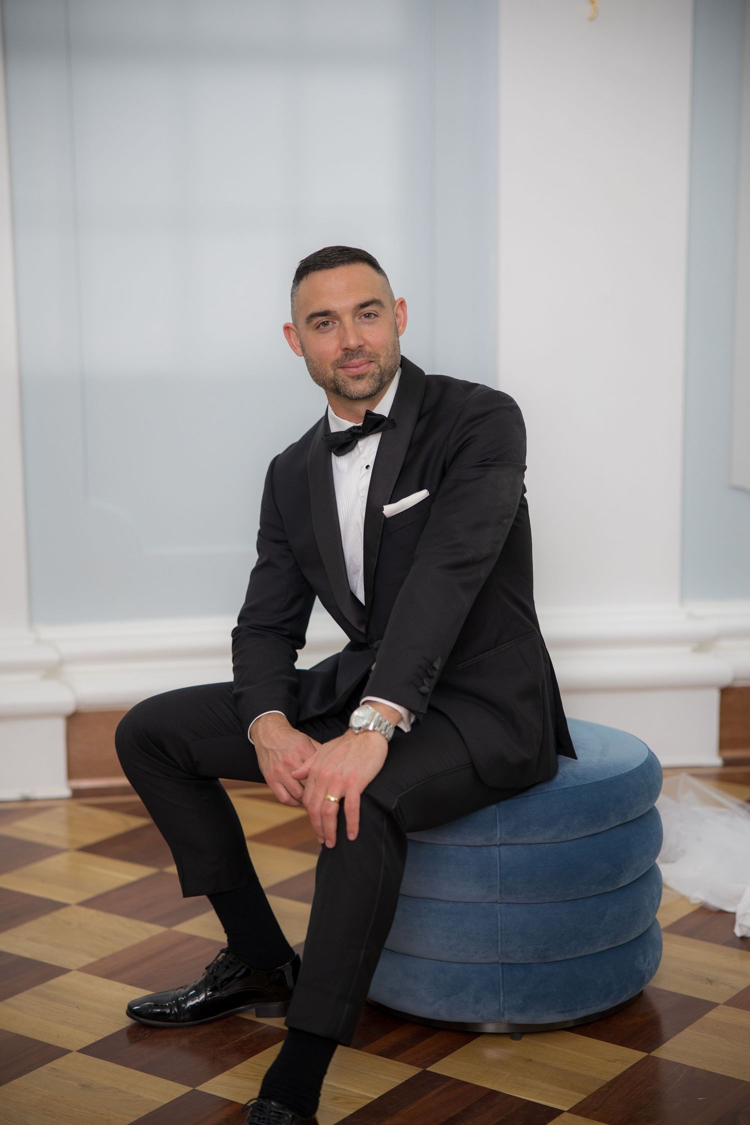 Dapper groom in black tuxedo sits confidently on blue velvet ottoman on checkered floor at wedding venue.