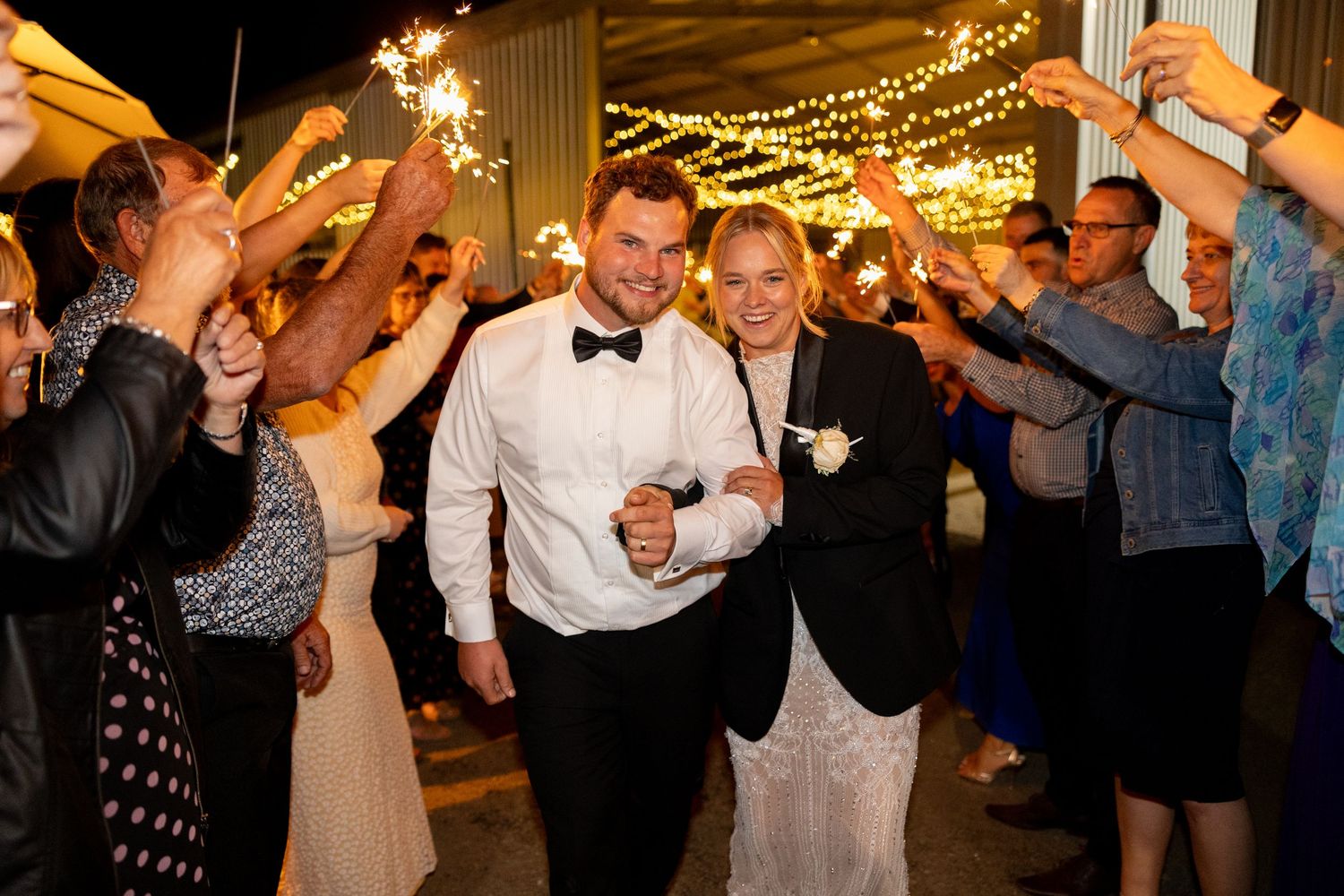 Newlyweds walk through a sparkler tunnel surrounded by cheering guests at nighttime wedding reception.