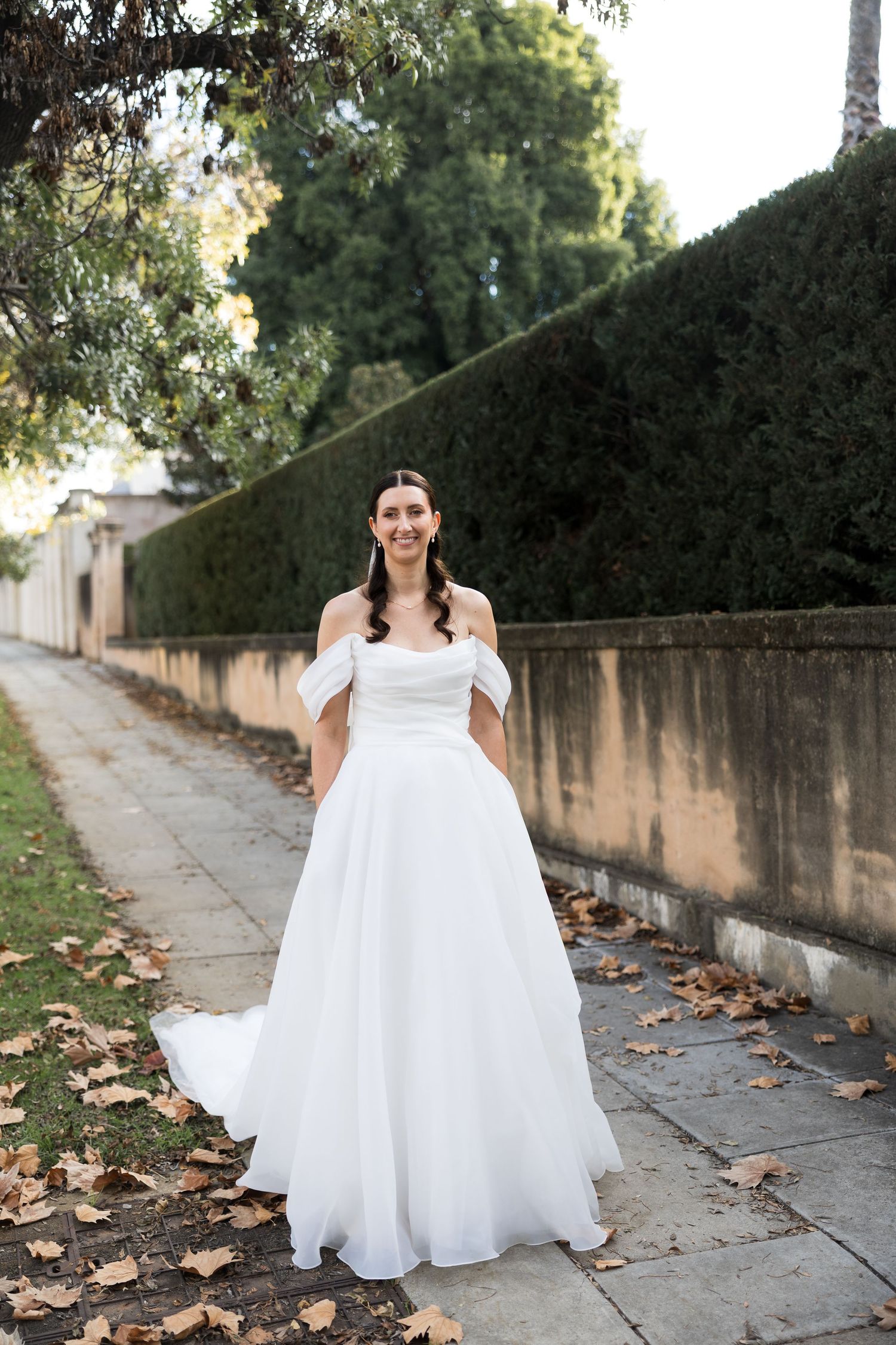 Bride in off-shoulder white ballgown wedding dress standing on a tree-lined sidewalk with autumn leaves.