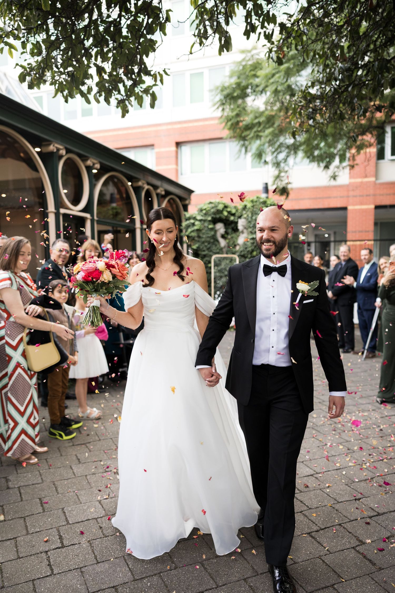 Bride and groom walk hand-in-hand through confetti shower, guests cheering, bride in off-shoulder gown holding bouquet.