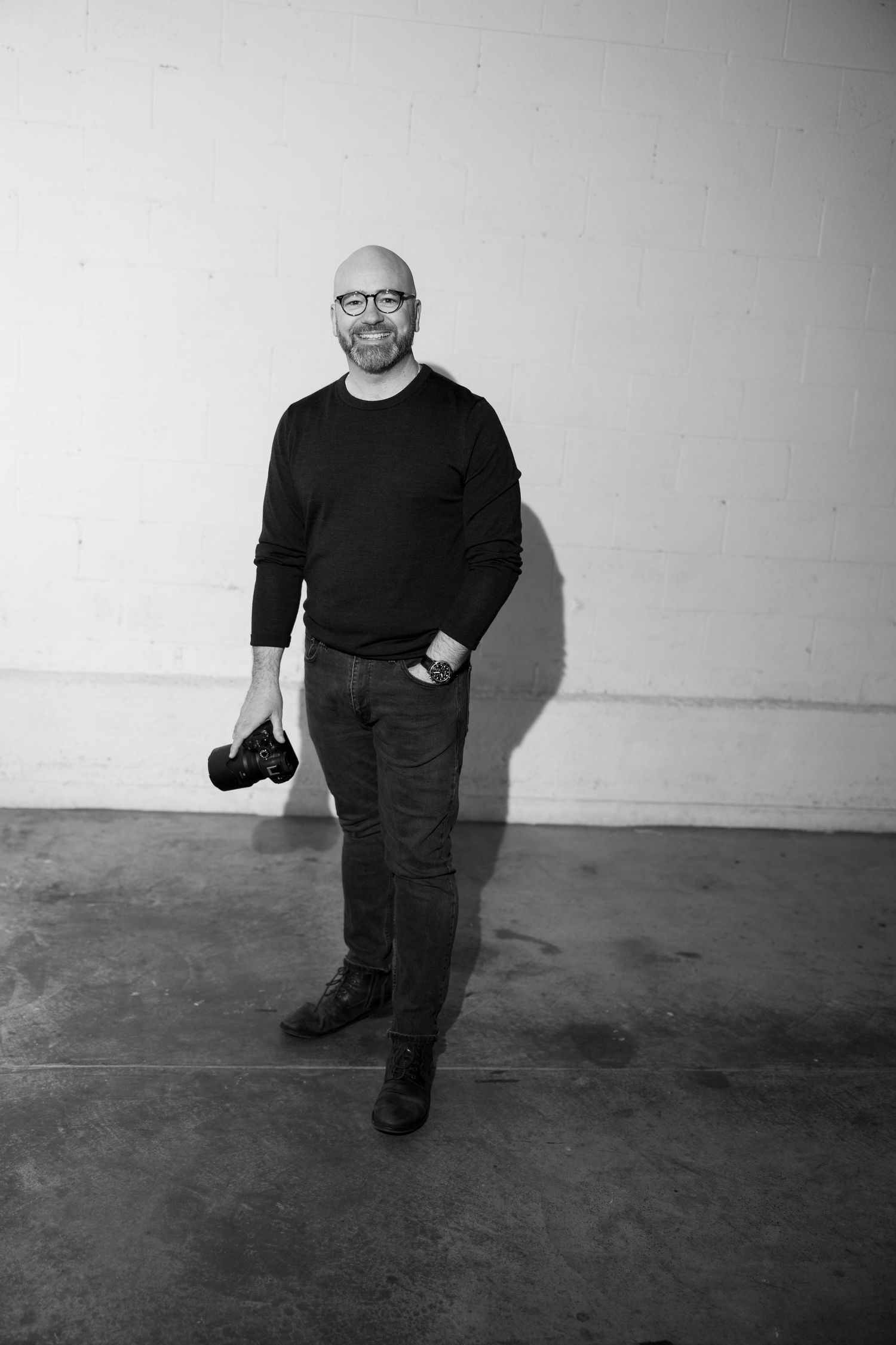 Bald man with glasses holding a camera, smiling in a black sweater and jeans against a white brick wall.