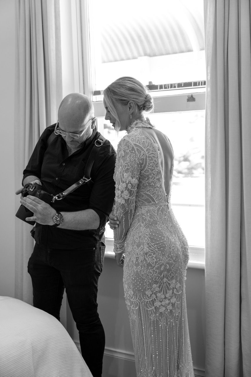 Photographer reviews camera with bride in lace wedding gown by window in black and white photo.