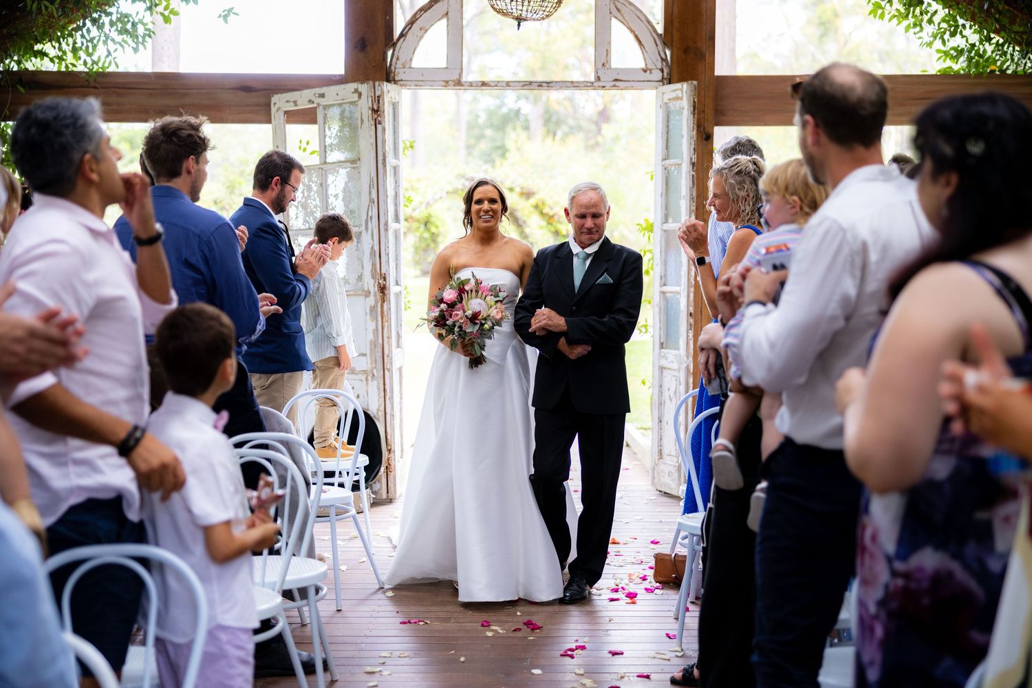 A bride walks down the aisle escorted by an older gentleman as guests watch in a rustic chapel venue.