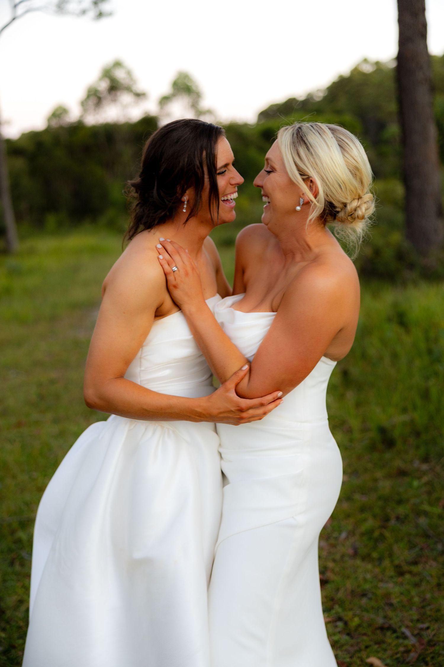 Two brides in white strapless wedding gowns smile at each other while embracing in an outdoor setting.