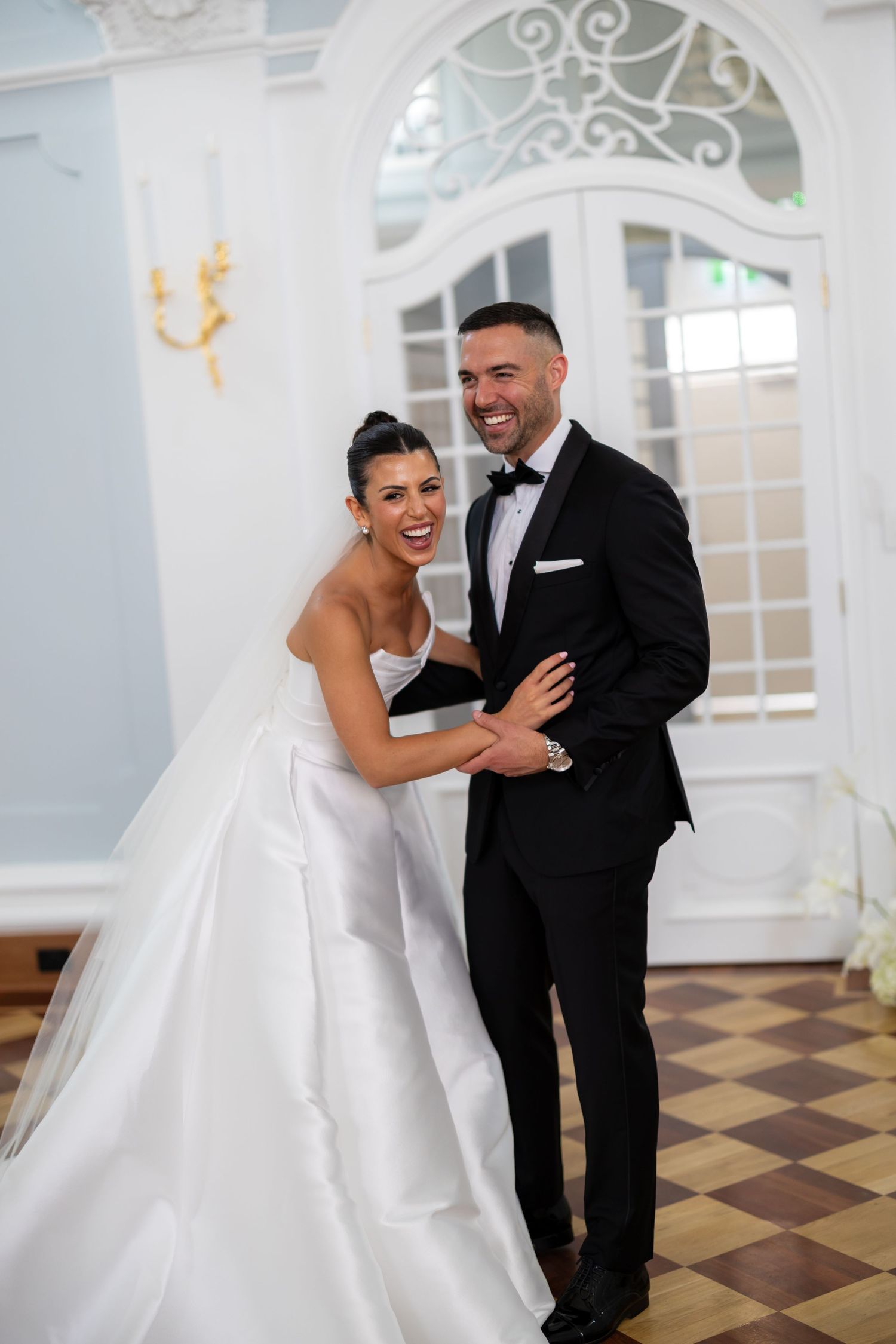 Smiling bride and groom laugh together in elegant ballroom with ornate white arched doors.