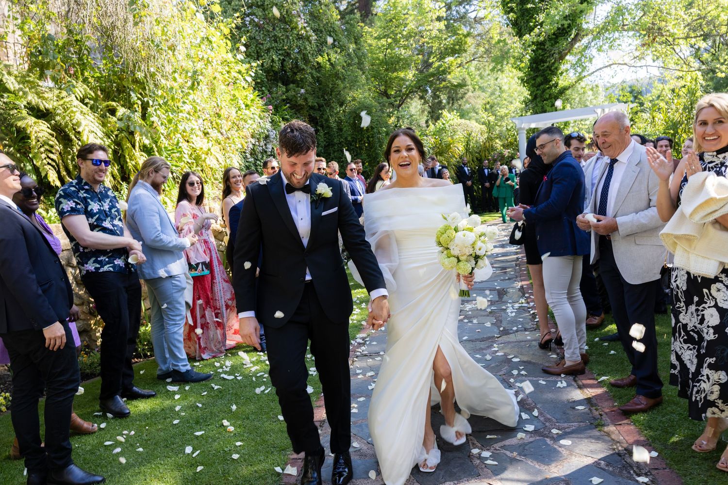 Bride in white off-shoulder gown and groom in tuxedo walking through confetti at outdoor ceremony.