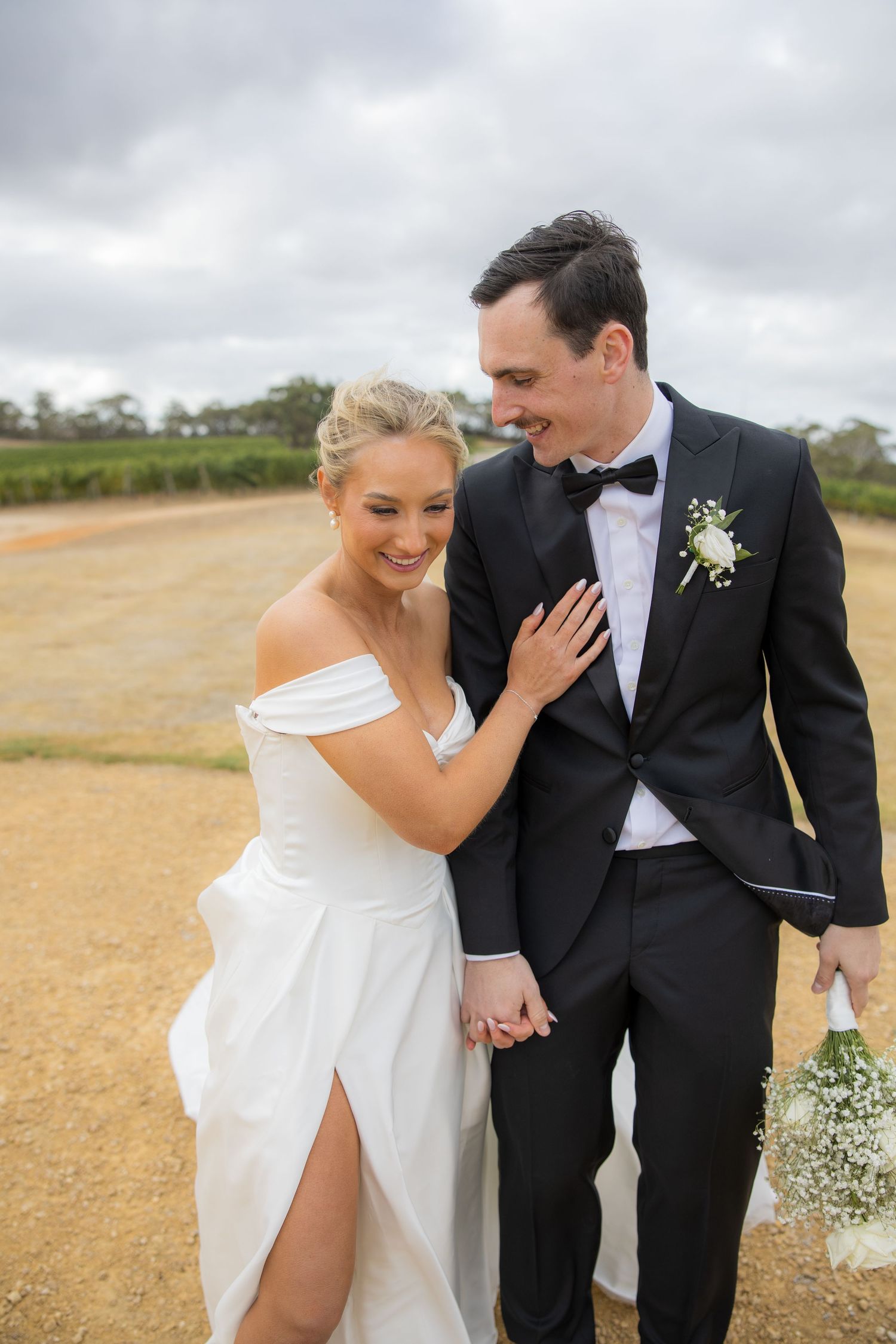Smiling bride in off-shoulder white gown holds grooms hand outdoors at vineyard wedding.